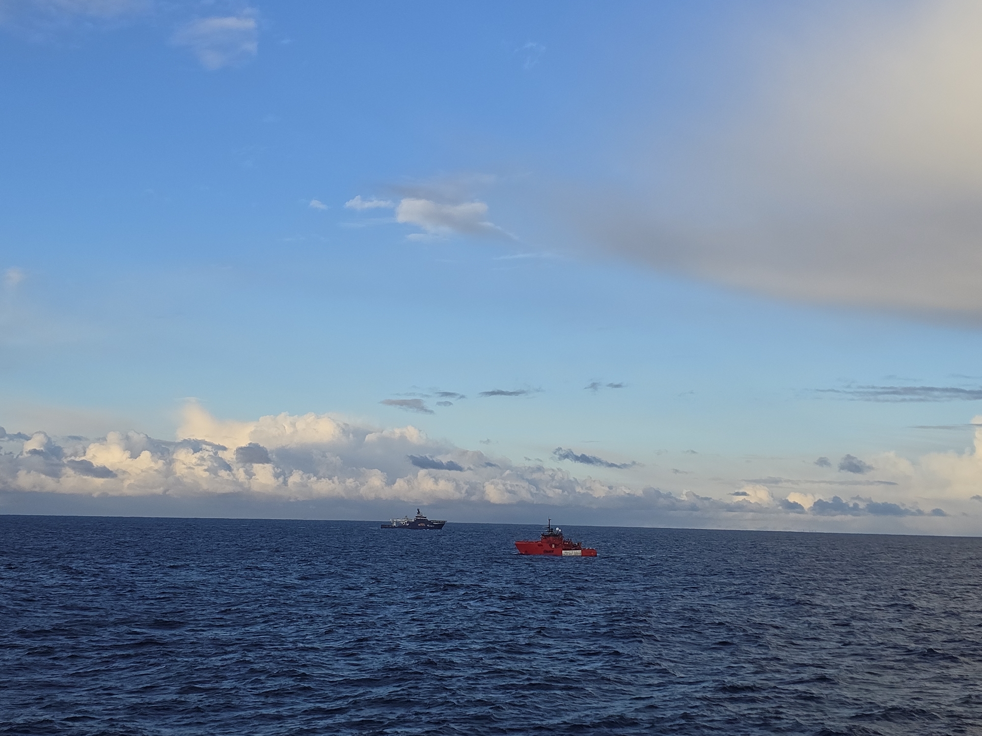 The image depicts two boats on a dark blue ocean under a sky with scattered clouds. One boat is red and has "COASTAL EVAL" printed on its side. The other boat is dark in color and situated further in the distance. There are no identifiable people visible in the image. The location appears to be on the open sea.