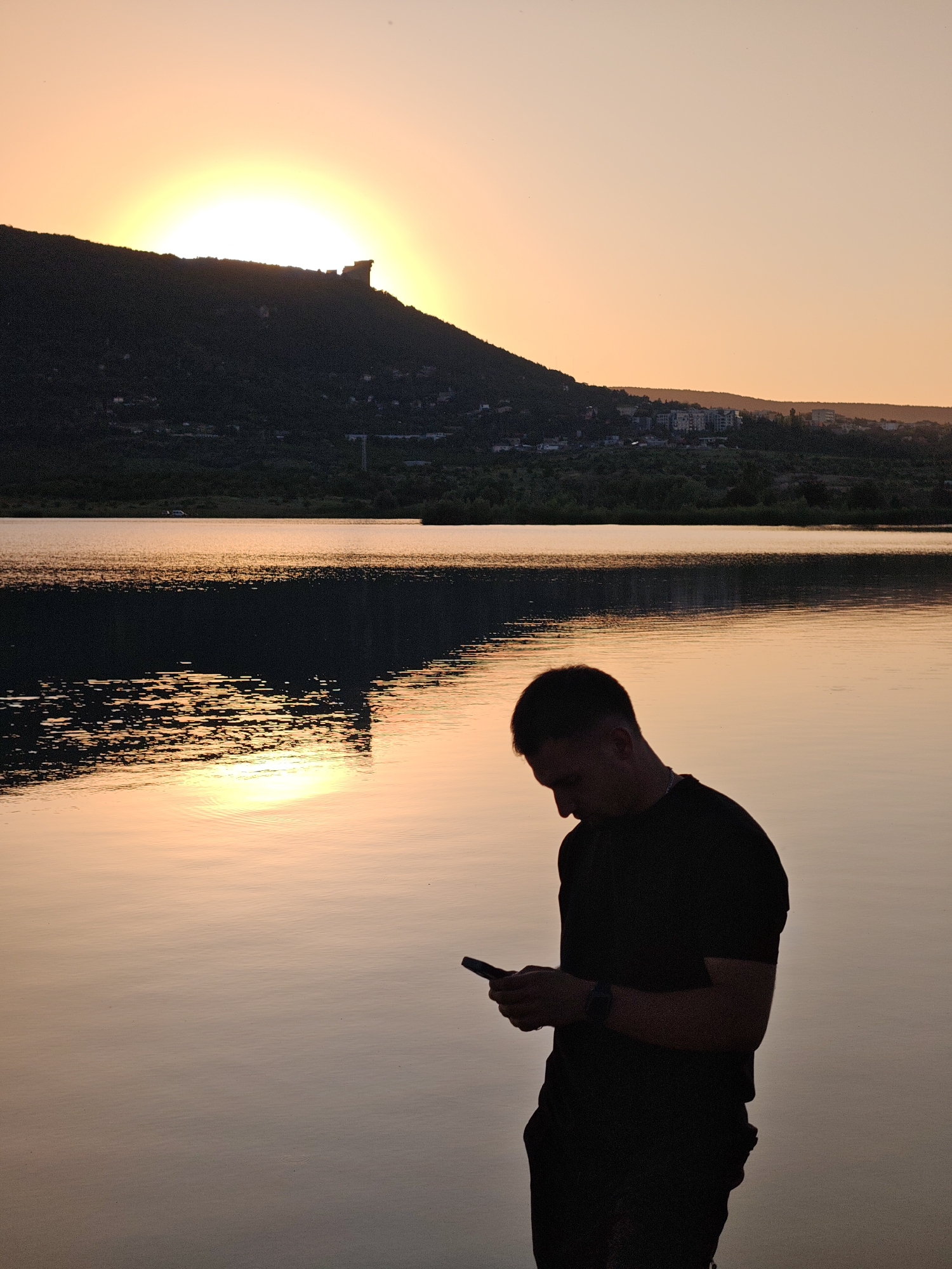The image depicts a silhouette of a young man standing by a body of water at sunset. He is looking down at his phone. In the background, a large, dark hill is visible, with the setting sun peeking over its peak. The sky is a gradient of orange and yellow. The water reflects the colors of the sky and the silhouette of the hill. The location is by the lake in the city of Shumen, Bulgaria.