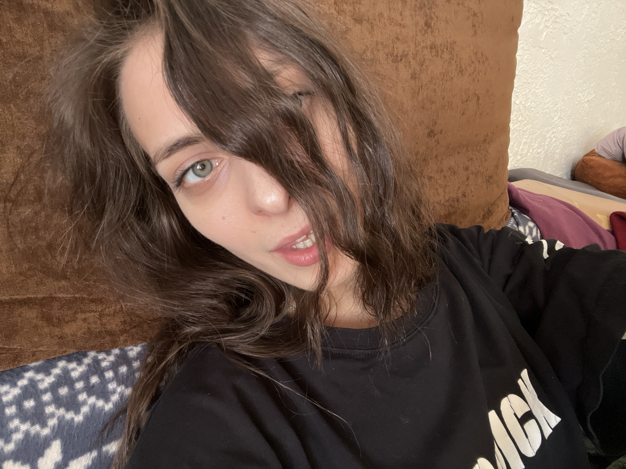 A woman with long, brown hair that partially obscures her face, is the primary subject of this indoor selfie. She has light skin, green eyes, and is wearing a black sweatshirt with white lettering. The background is composed of a brown, textured cushion or headboard, and a glimpse of a patterned blue and white surface, possibly a blanket or bedspread. The environment suggests a casual, relaxed setting, likely a home interior. There's no indication of the specific location, or city of Krasnoyarsk. No information regarding specific objects, actions, or interactions beyond the selfie.