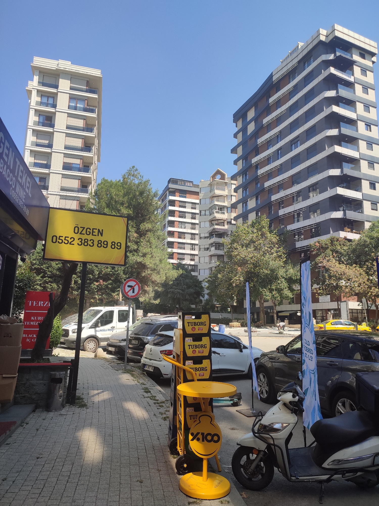 This is a street-level, eye-level medium shot captured on a bright, sunny day, likely in Kadıköy, Turkey, judging by the street signs and building architecture. The scene depicts a sidewalk lined with trees, with several modern apartment buildings in the background.
Dominating the left side of the frame is a large yellow advertising sign for "ÖZGEN" with the phone number "0552 383 89 89". To the left of this sign, a dark blue sign with white lettering, partially visible, reads "GEN TEKEL" and "BACCO WINE". Below the yellow sign, on a grey wall, is a red sign that reads "TERZA". Further down the wall, smaller signs indicate "KURU TEMİZ" and "BLACK IC ÇI".
On the sidewalk, several vehicles are parked. A white commercial van, labeled "HALIMOĞLU İNŞAAT", is prominent. Adjacent to it are a dark colored SUV and a white hatchback car.
A yellow table with a small umbrella and a stand advertising "TUBO R G" and "X100" is positioned on the sidewalk. A white scooter is parked to the right of this display. Two blue and white flags are also present on the sidewalk.
The environment is a typical urban street scene with paved sidewalks and roads. The buildings are contemporary in design, featuring balconies and large windows. The trees are lush and green, suggesting a temperate climate. There are no people clearly visible engaging in any specific actions, though the presence of parked vehicles and commercial signage implies a public commercial area. The lighting indicates it's daytime, with shadows cast on the sidewalk.