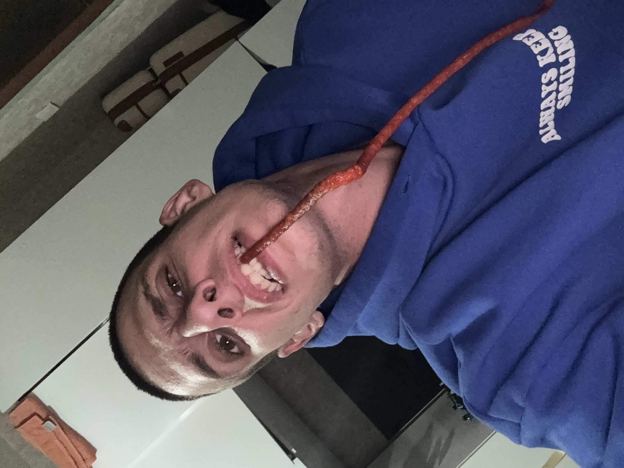 A rotated, close-up, medium shot captures a young adult male in an indoor setting. He wears a bright blue hooded sweatshirt, featuring white text that reads "ALWAYS KEEP SMILING" on the right sleeve/shoulder. The man looks directly into the camera with wide, dark brown eyes and an intense, perhaps playful or humorous expression. His mouth is slightly open, revealing his upper teeth. A long, thin, red, textured stick-like object, resembling a snack or food item, is positioned in front of his face, extending from near his mouth/nose area upwards towards the upper right of the frame.
The background, partially visible and slightly out of focus, shows elements of an indoor environment. In the upper left, two light-colored, rectangular objects with dark straps, possibly small bags or cases, are placed on a shelf or ledge. Below the man's head, an orange folded towel or cloth is partially visible on a light-colored surface. The lighting suggests an interior space, likely a room or kitchen. This scene takes place in Sofia, Bulgaria.
