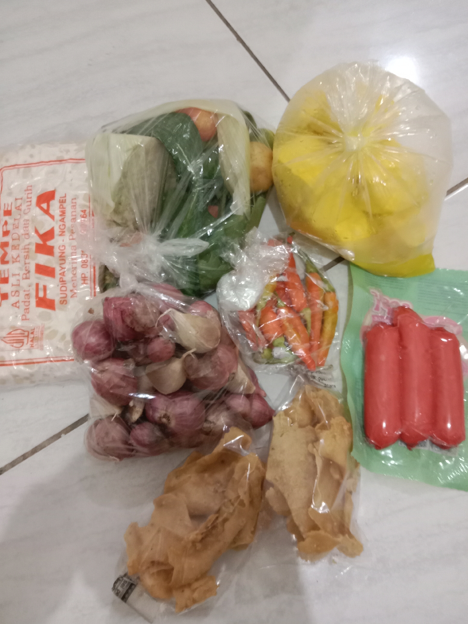 A collection of groceries is arranged on a tiled floor, likely in Brangsong, Indonesia. The items include a package of tempeh, a bag of shallots and garlic, a bag of chilies, a bag of unidentified yellow fruit, a bag of snacks, and a package of sausages. The lighting suggests an indoor setting, possibly a kitchen or pantry. The floor is made of light-colored tiles, with visible grout lines.