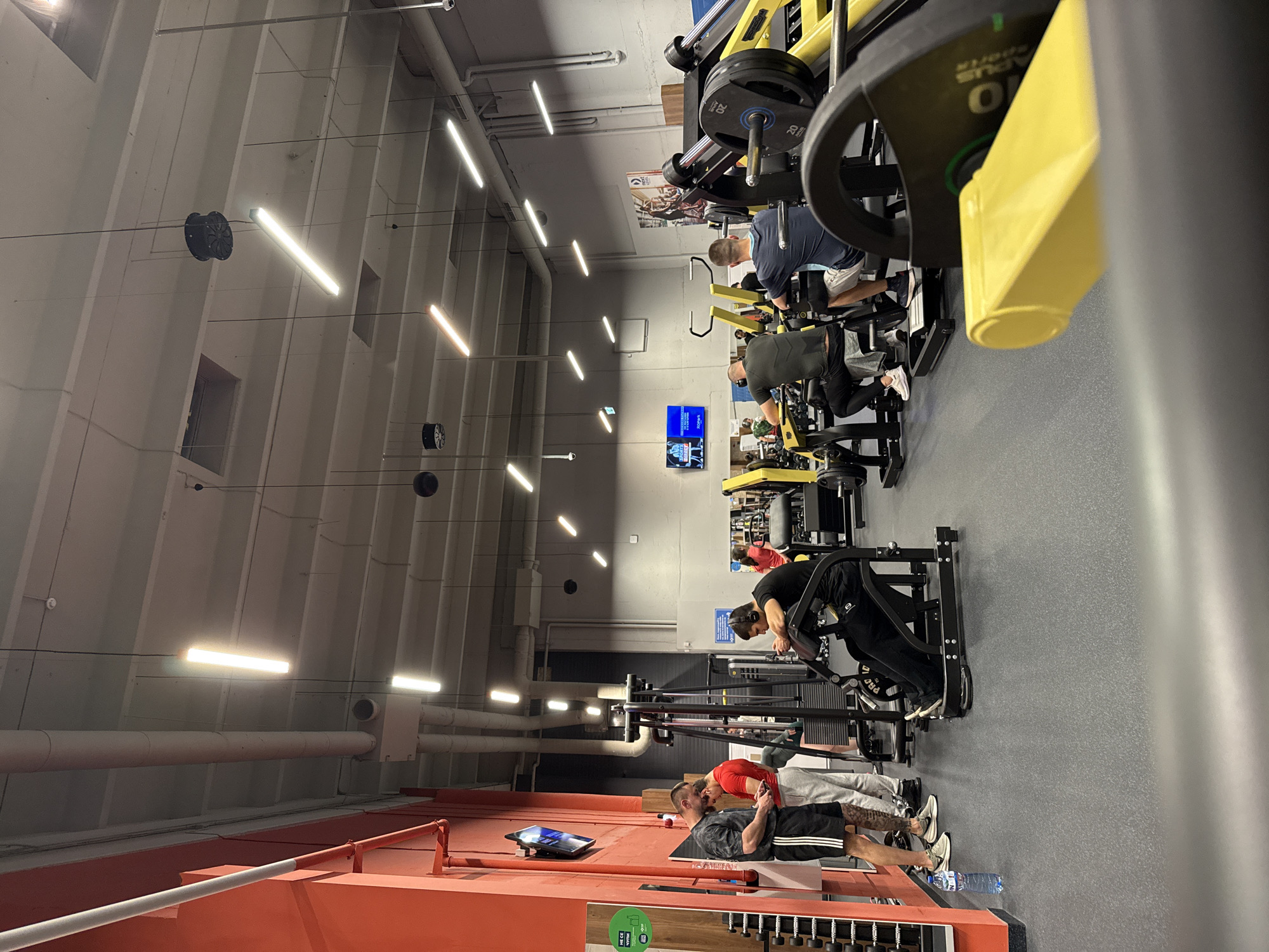 The image depicts the interior of a gym. Several individuals are engaged in workouts on various exercise machines. The gym has a modern aesthetic with overhead fluorescent lighting, dark grey flooring, and red wall accents near the bottom of the frame. The gym appears to be well-equipped with a variety of weight machines. The location is presumably in Sofia, Bulgaria.
