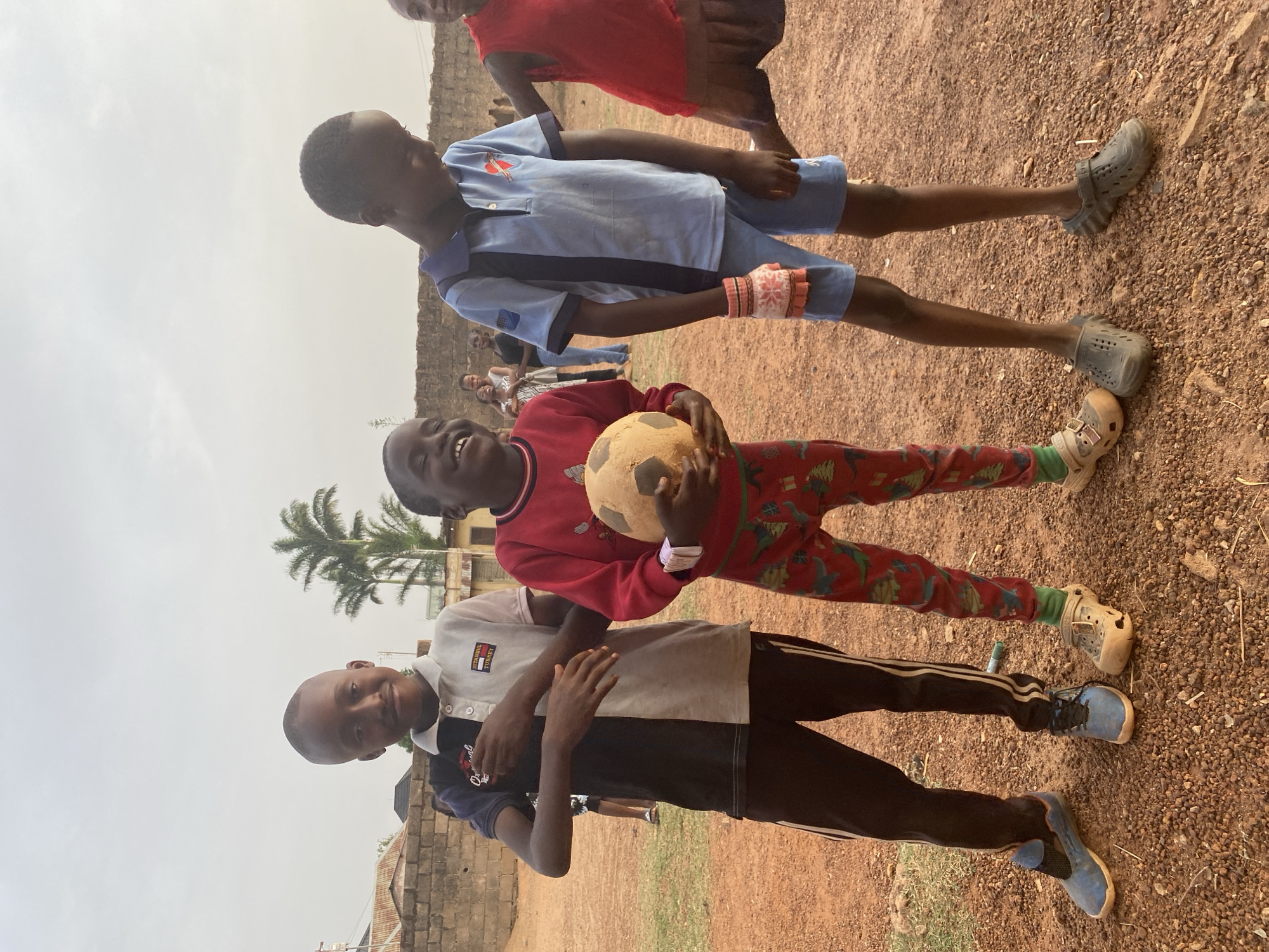 The image captures three young boys smiling brightly as they pose for a photo outdoors. They stand side-by-side on reddish-brown dirt or gravel ground, suggesting an unpaved area like a community space or yard.
In the center, a boy with a broad, joyful smile holds a worn, light-colored football with black markings. He wears a red long-sleeved top and red trousers patterned with green figures, along with light-colored Crocs-style shoes and green socks. To his left (viewer's right), another boy with a pleasant smile is dressed in a two-toned polo shirt—light gray on top and black on the bottom—paired with black track pants featuring white stripes. He wears blue sneakers, and text on his shirt reads "ISTANBUL TURKEY" and "Original". To the central boy's right (viewer's left), a third boy, also smiling, wears a light blue polo shirt and matching light blue shorts, with dark gray Crocs-style shoes. He has a patterned, multi-colored wristband on his left arm.
The background features low-rise buildings or walls made of reddish brick or concrete blocks. A palm tree is visible behind the central boy. The sky above is bright but appears hazy or overcast. Several other blurred figures, both children and adults, can be seen in the mid-ground, indicating a larger informal gathering or activity. The overall atmosphere is one of happiness and community, likely during playtime in a warm climate.