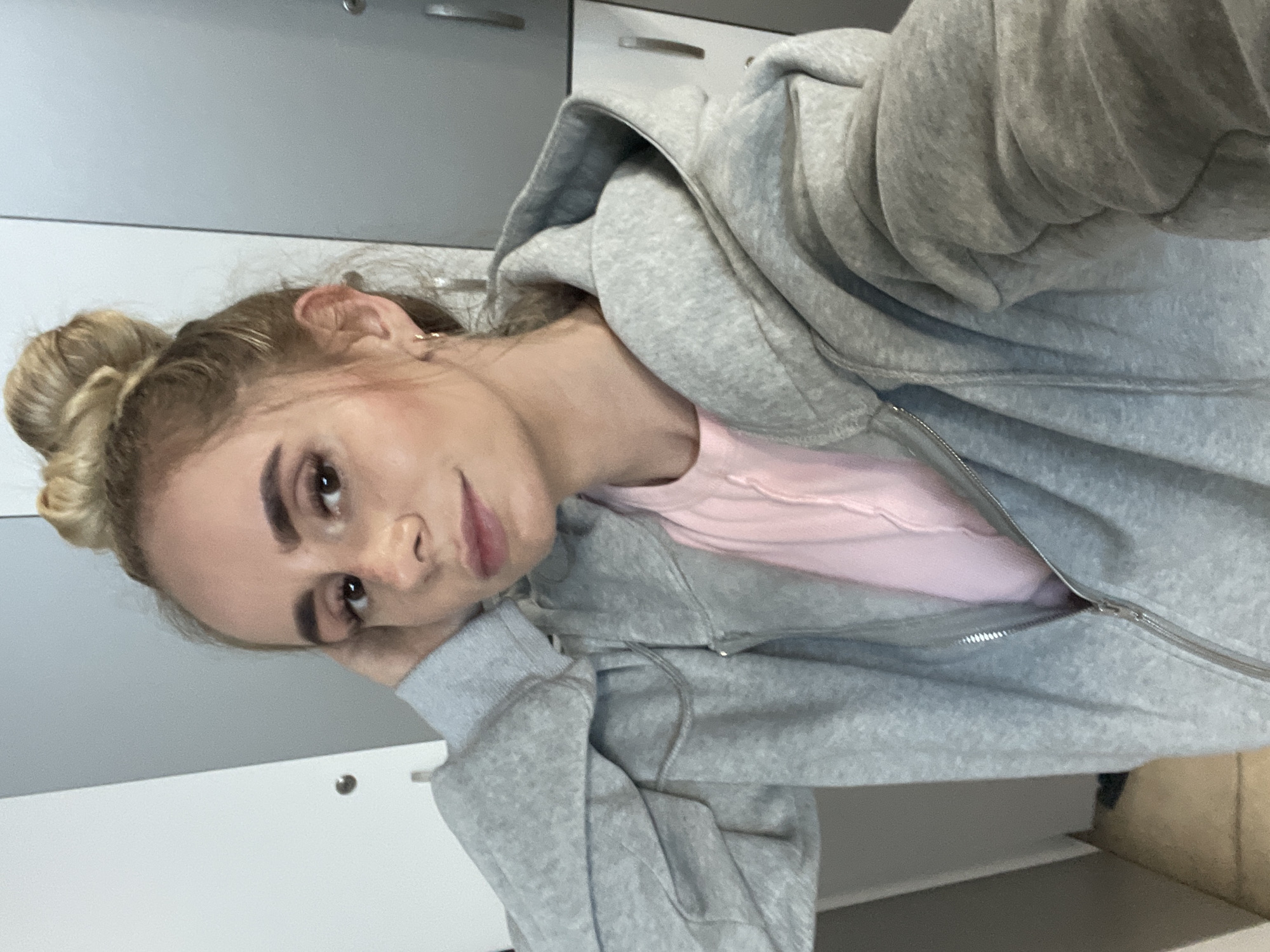 The image shows a young woman with blonde hair styled in a bun, wearing a light pink t-shirt and a grey zip-up hoodie. She is looking towards the camera with her right hand resting on her head. She is in a locker room setting, with grey lockers visible in the background. The lighting suggests an indoor environment. There is no indication of the specific location within Vladivostok, Russia.