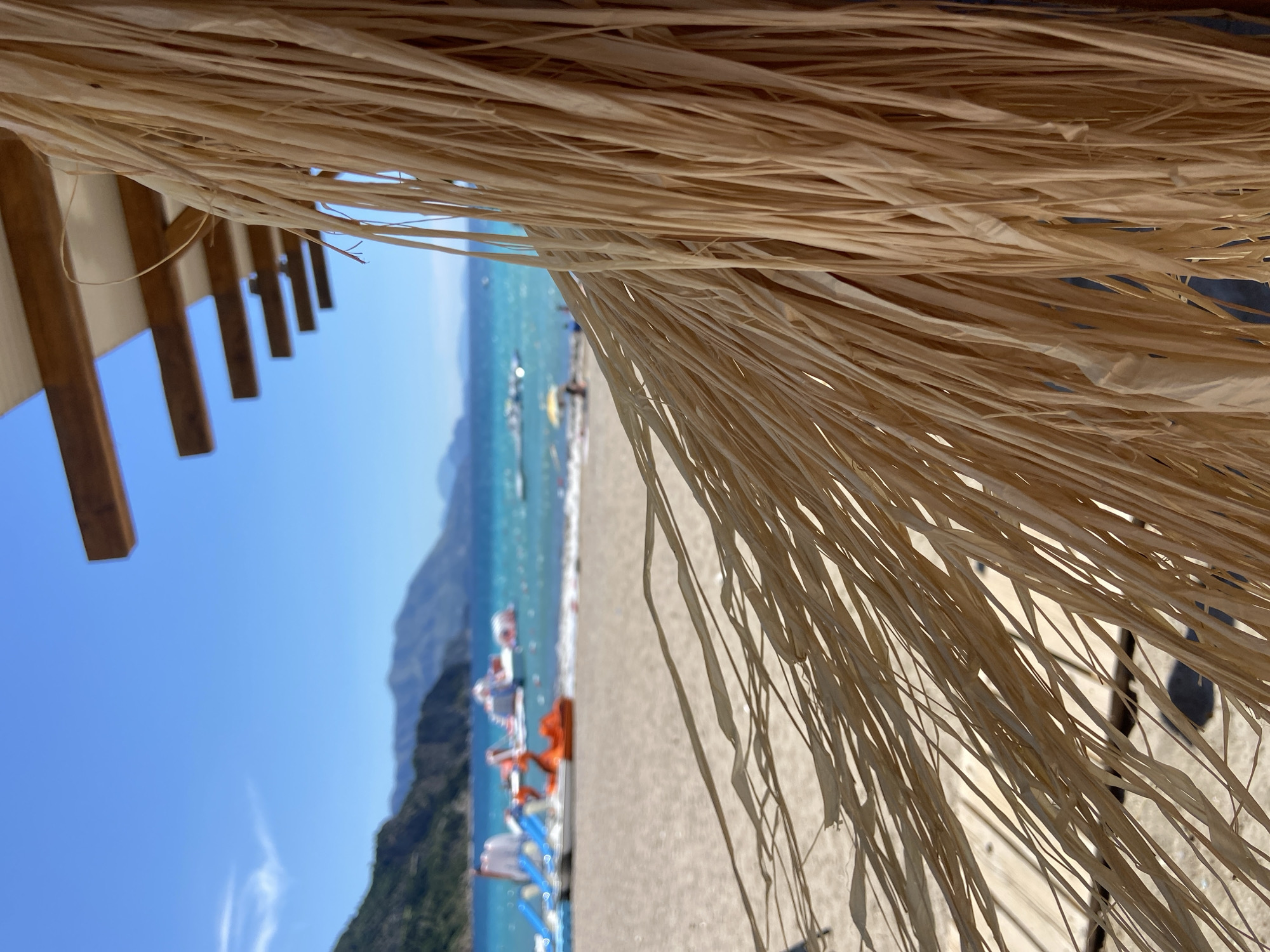 This is a close-up, low-angle, eye-level shot of a thatched roof overhanging a beach in Prijevor, Montenegro. The thatched material, made of dried grass, is in the foreground, obscuring the view of the beach and sea. The blurred background reveals a sandy beach with several colorful watercraft, including paddle boats and what appear to be inflatable water toys, on the turquoise sea. In the distance, rolling green mountains meet the bright blue sky. The scene is bathed in sunlight, creating a bright and cheerful atmosphere.