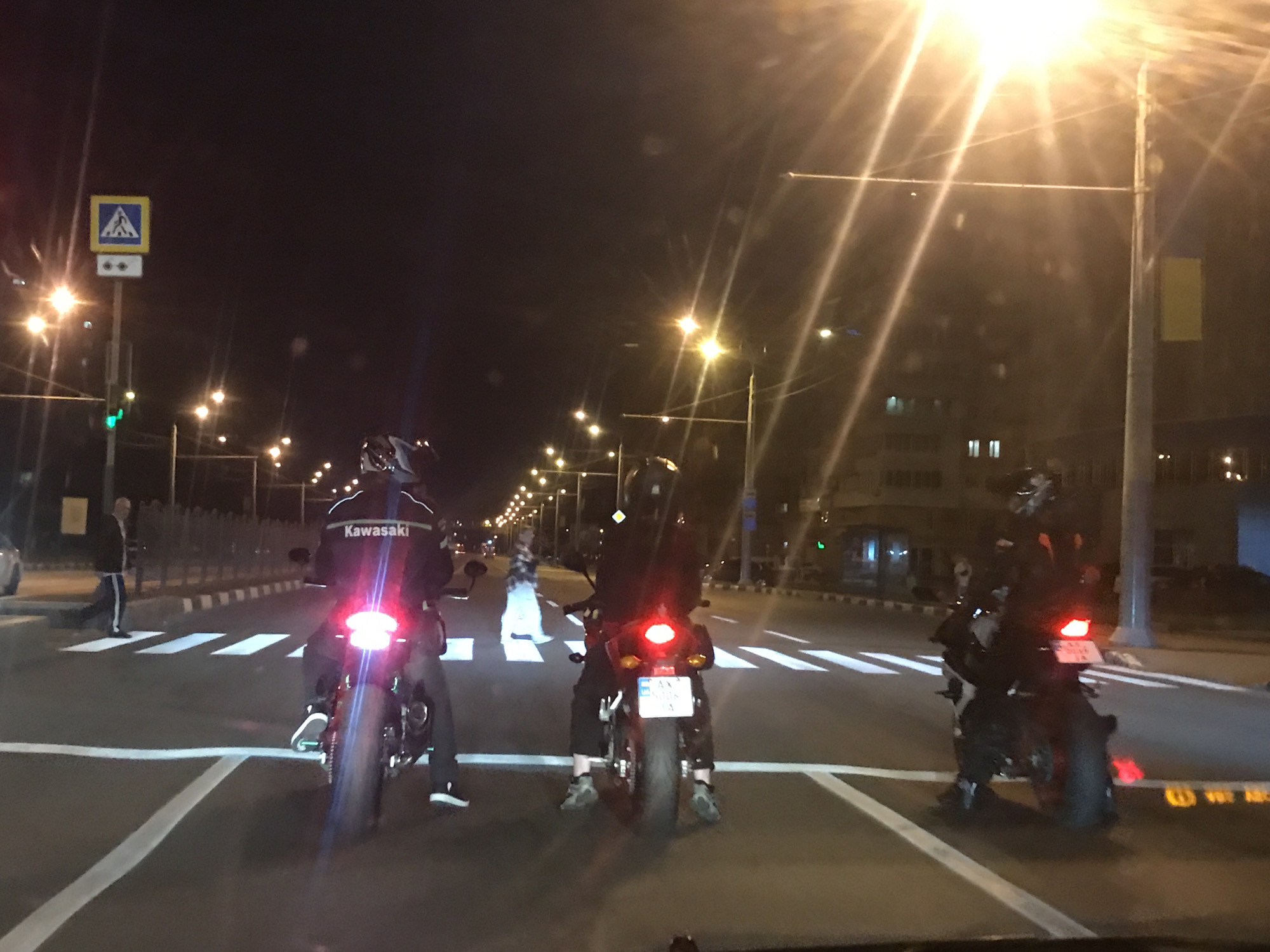 The image depicts a night scene on a city street, viewed from inside a vehicle. Three motorcyclists are stopped side-by-side at a pedestrian crossing, their motorcycles' red taillights illuminated. The motorcyclist on the far left wears a jacket with "Kawasaki" written in white across the back. The middle motorcycle displays a partially visible license plate that appears to be Armenian, starting with "AX" followed by numbers like "0006".
The setting is characterized by darkness punctuated by numerous bright streetlights that create pronounced light streaks and lens flares across the frame. Buildings with some illuminated windows are visible in the background, suggesting an urban environment. A pedestrian is seen walking across the marked white stripes of the crosswalk in the mid-ground, moving from left to right.
To the left, a blue square sign with a white triangular inset showing a black pedestrian symbol marks the crossing. Below it, a white rectangular panel with two dark circular elements is attached to the pole. White road markings, including stop lines and pedestrian crossing stripes, are clearly visible on the asphalt. The overall activity involves vehicles stopped at a pedestrian crossing at night.