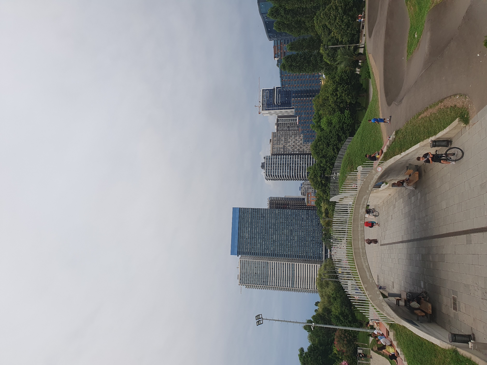 This image captures a view of a modern urban park in Batumi, Georgia, with a cityscape as the backdrop. In the foreground, a pedestrian walkway curves under an arched concrete structure, leading to a paved area with benches. Several people are visible, some sitting on benches and steps, while others walk or engage in recreational activities. One person is riding a bicycle, and another is on a scooter. To the right, a dirt track with undulating mounds, likely a pump track, suggests a space for cycling and skateboarding.
Beyond the park, a line of lush green trees forms a natural barrier between the park and the city. Dominating the skyline are several tall, contemporary buildings, characteristic of modern urban development. One prominent skyscraper features a blue-tinted upper section, while others display a mix of glass facades and varied architectural designs. A construction crane is visible near one of the buildings, indicating ongoing development. The sky is overcast, casting a soft, diffused light over the scene. The overall impression is one of a vibrant, public space seamlessly integrated into a developing urban environment.