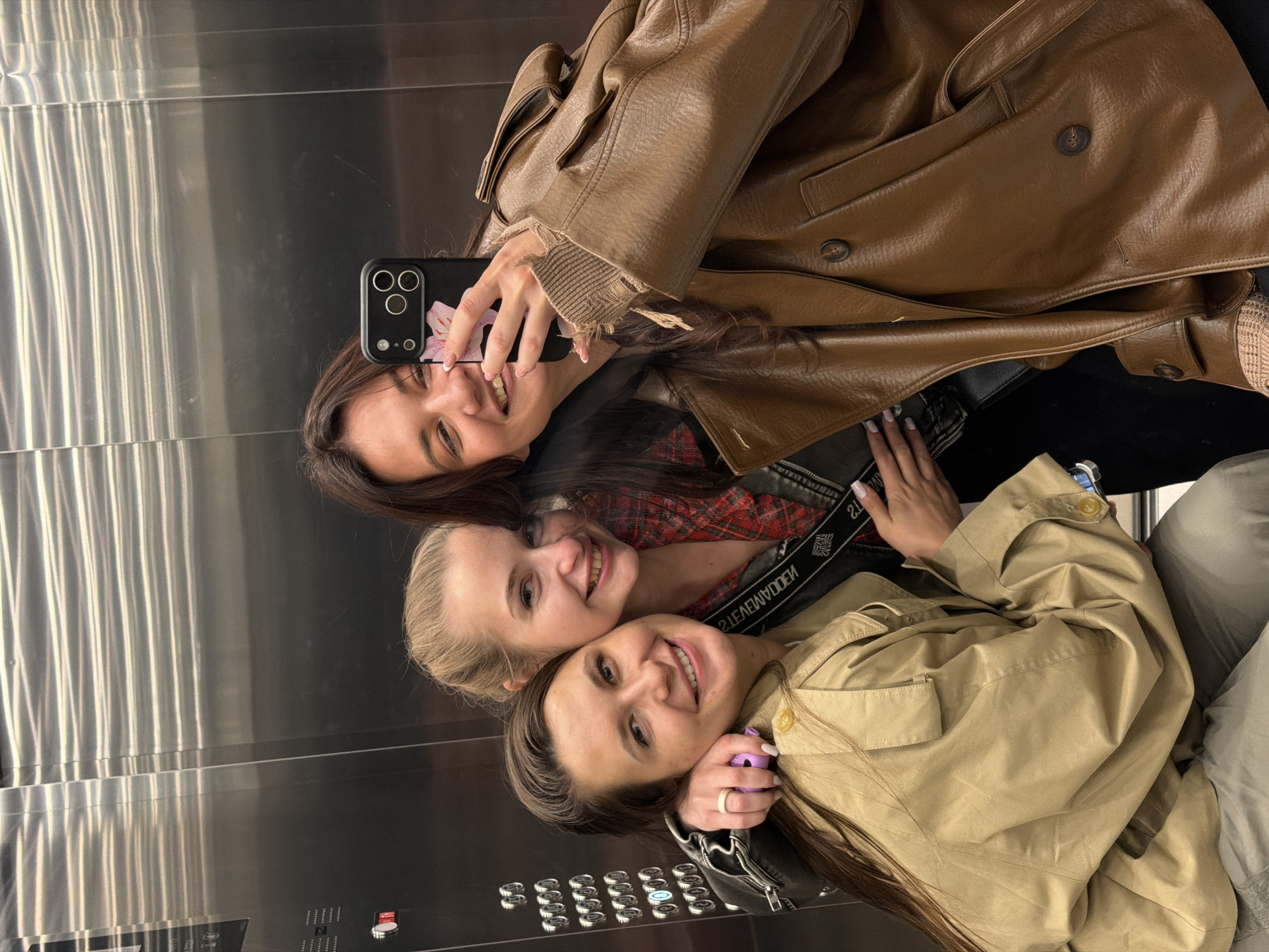 Three young women are taking a selfie in an elevator with a mirrored wall. The woman at the top is holding a smartphone with a floral design on the case. She is wearing a brown leather jacket. The woman in the middle has blonde hair and is wearing a red and black checkered shirt. The woman at the bottom is wearing a beige trench coat. All three women are smiling and looking at the camera. The elevator panel shows a series of buttons, and the surrounding environment appears to be a modern, urban setting. There is no visible location or clue that indicates they are in Belgrade, Serbia.