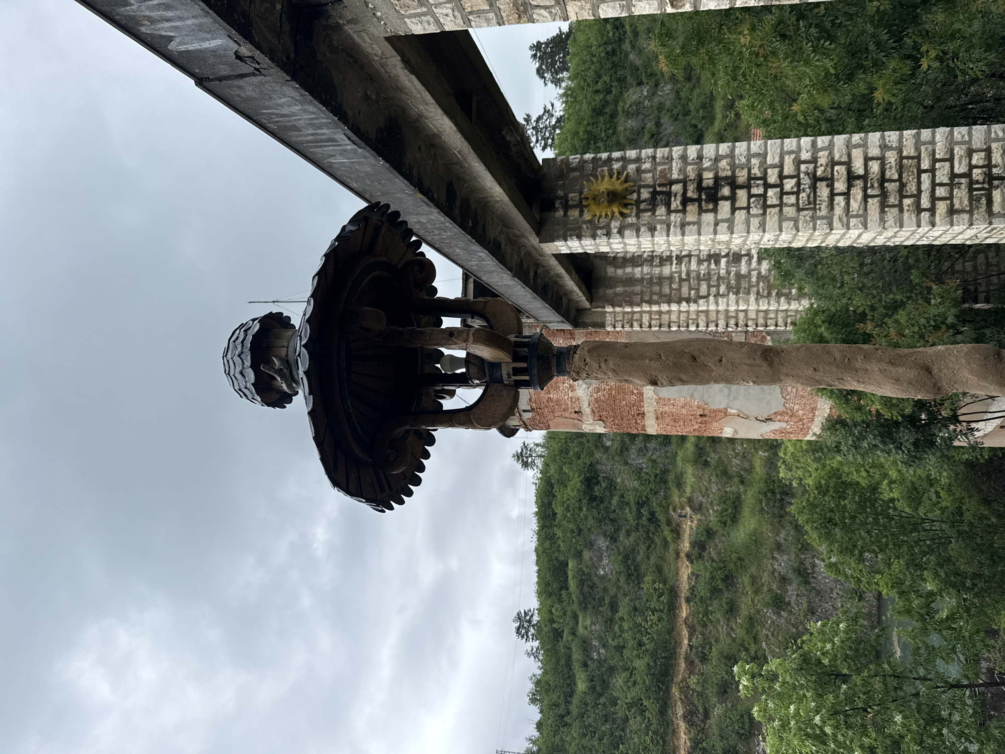 This is an upward-facing shot of the monument of "The Sun" in the city of Lukovit, Bulgaria. The statue sits under the remains of a stone structure, the column of which is constructed with gray and red brick. On the column behind the statue is a sun decoration. The background includes a forested hillside and a cloudy sky.