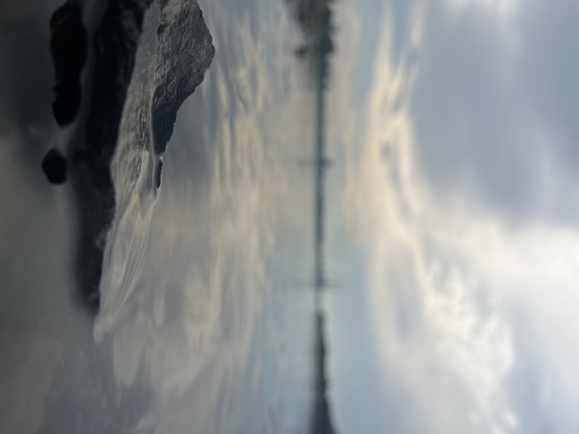 The image depicts a close-up view of a rock emerging from the surface of a body of water. The water is calm and reflects the cloudy sky above. In the distance, the blurred outline of a cityscape is visible, suggesting an urban or industrial setting. The focus is on the textured surface of the rock and the gentle ripples it creates in the water. The overall impression is one of quietude and a blend of natural elements with a distant human presence. The image is too abstract to identify a specific location within Kemerovo, Russia.