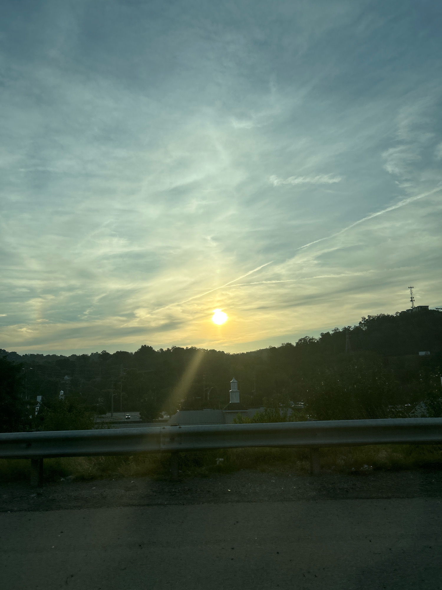 The image captures a view of a hilly landscape with a sunset in Collier Township, United States of America. The sun, a bright yellow orb, is positioned towards the center-right of the frame, casting a warm glow through a hazy sky. Wispy clouds, tinged with yellow and blue, stretch across the sky, with visible contrails from aircraft. In the middle ground, a densely wooded hillside is visible, with scattered buildings and structures peeking through the foliage. A white steeple, possibly belonging to a church or mosque, stands out against the green. A metallic guardrail runs horizontally across the lower portion of the image, separating a paved road from the embankment beyond. The road surface in the foreground is dark and appears to be asphalt. A light-colored vehicle is visible on a road in the distance, traversing the hillside. The overall scene depicts a natural and semi-urban environment bathed in the soft light of sunset.