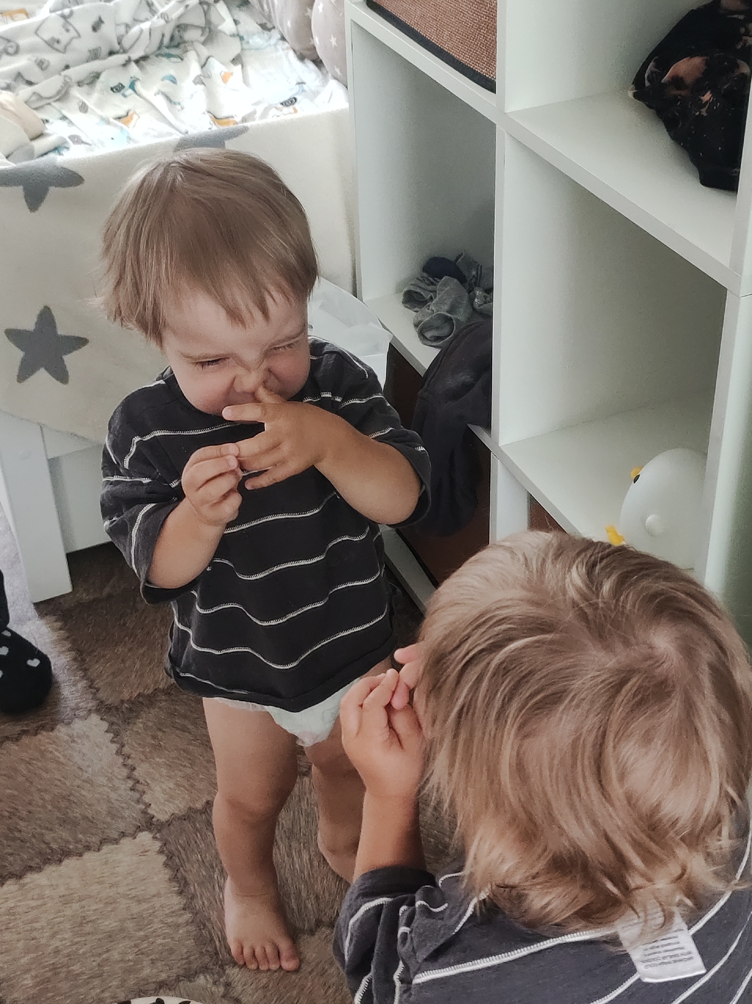 The image depicts two young children indoors, with one child in the foreground facing away from the camera and the other child in the middle ground facing towards the camera. The child in the middle ground is wearing a striped t-shirt and a diaper, and is picking their nose. The child facing away has blonde hair and is wearing a striped shirt. The environment appears to be a child's room or play area, with a shelf unit containing various items, including clothing and a small white lamp. The floor is covered with puzzle-like mats. The overall lighting is natural, suggesting daytime. There is no visible indication of the location being in the city of Chaikovsky, Russia.