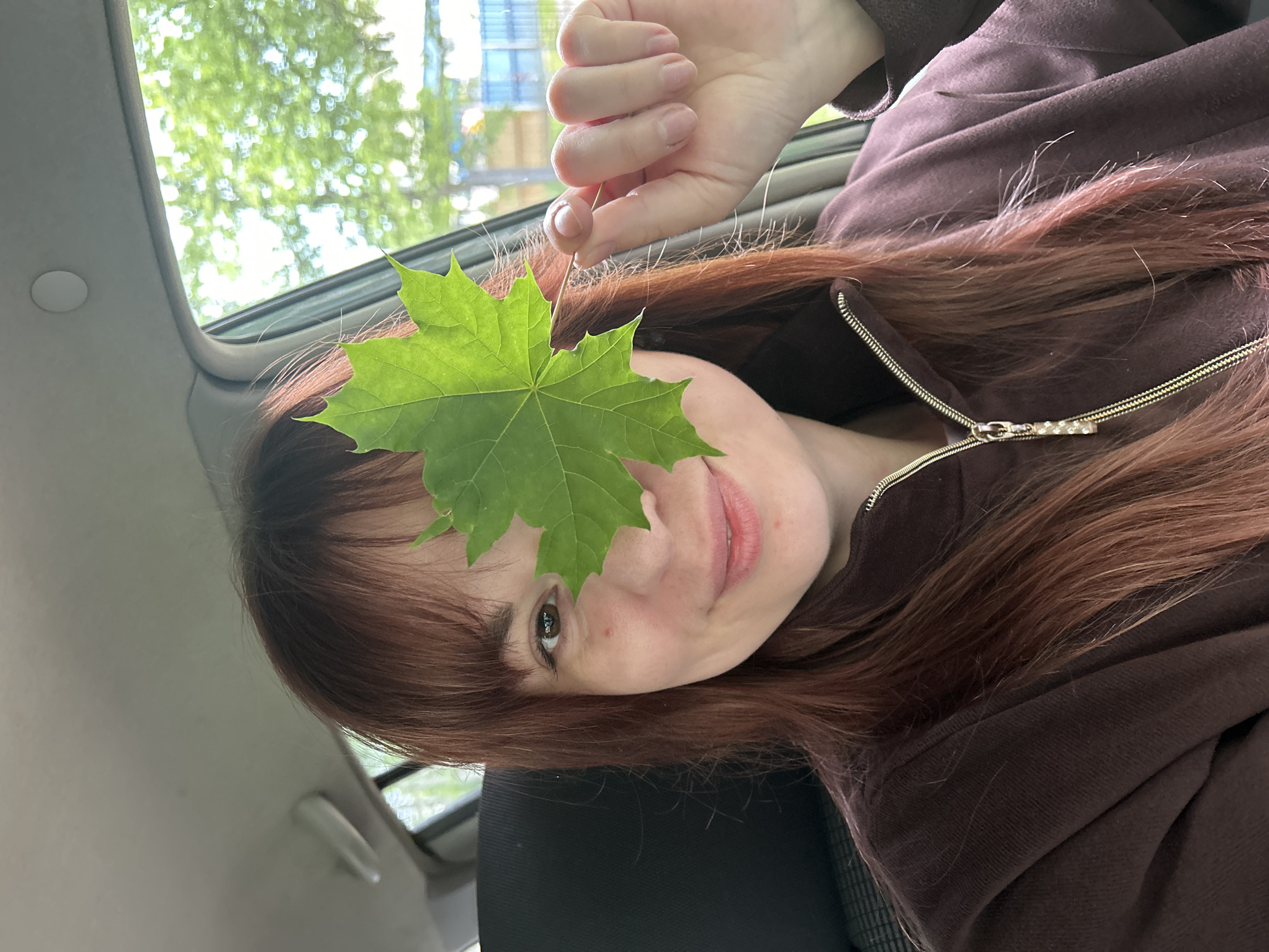 A close-up selfie captures a young woman inside a car, her image rotated 90 degrees clockwise. She has long, reddish-brown hair and is wearing a dark brown zippered hoodie or jacket with a golden, embellished zipper pull. Her right hand holds a bright green, maple-like leaf, positioning it to cover her left eye. Her visible right eye is dark, and she has a gentle, subtle smile. Light freckles are visible across her nose and cheek. Through the car's sunroof, located above her head in the rotated image, blurred green trees and bright daylight are visible, indicating it is daytime. The car interior is a light color. Her fingernails are neatly trimmed and unpainted. The overall mood is relaxed and playful. No visible text is present. The scene is set inside a car, likely in Chaikovsky, Russia.