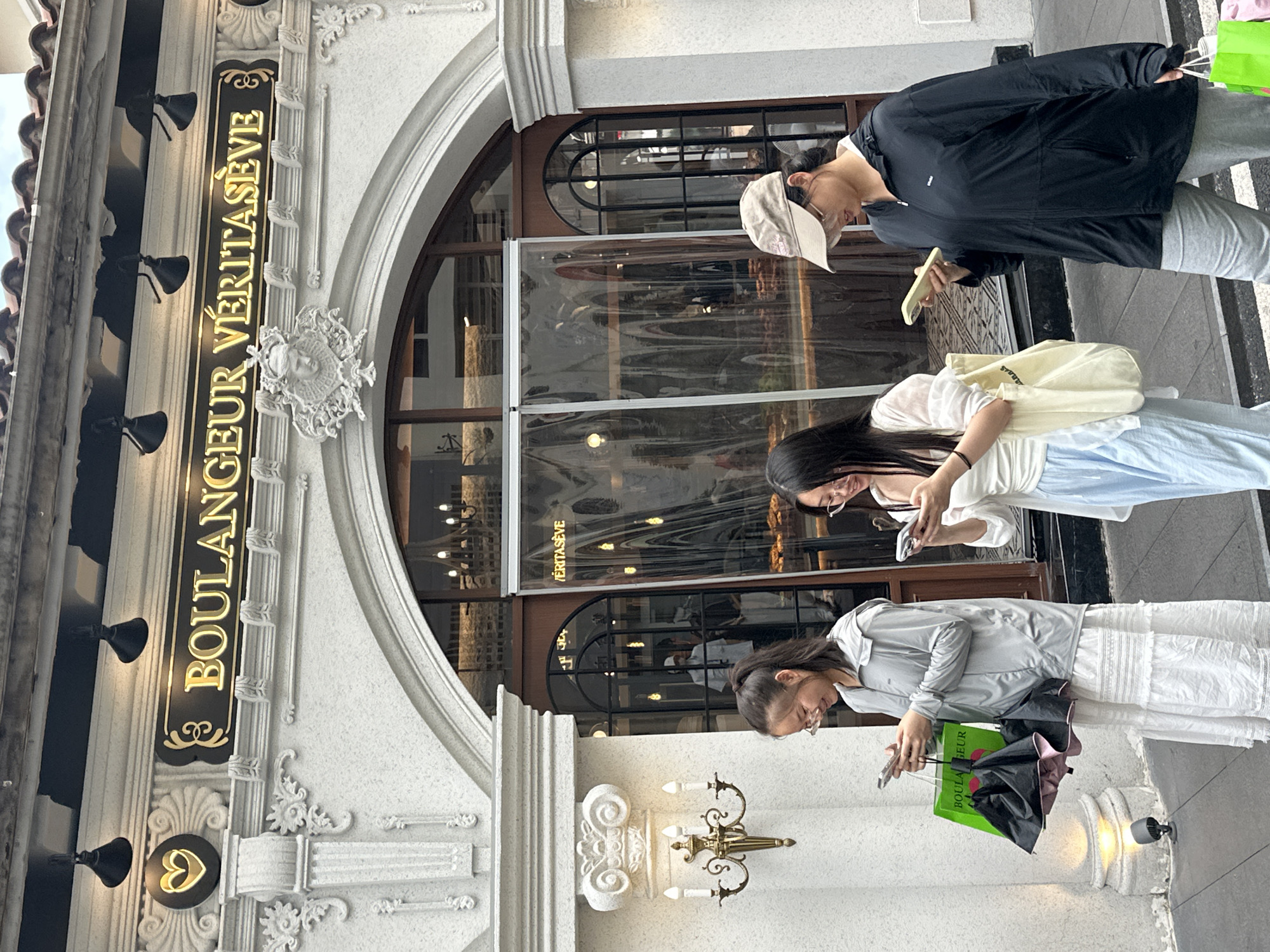 Three individuals are captured on a sidewalk in front of a bakery in the Chongqing High-tech Zone, China. The bakery's sign, illuminated in gold lettering on a black background, reads "BOULANGEUR VÉVITASEVE." The facade of the building is ornate and white, with architectural details and decorative lighting fixtures.
The person on the left, a young woman with her hair in a ponytail, wears glasses and a light gray hooded jacket over a white tiered dress. She holds a green shopping bag containing a closed black umbrella in her left hand and is looking down at her mobile phone, which she holds in her right hand.
The central figure, also a young woman wearing glasses, is dressed in a sheer white long-sleeved top with a white camisole underneath and light blue trousers. She carries a light-colored tote bag on her right shoulder and is looking down at a mobile phone in her left hand.
On the right, a woman wearing glasses and a beige baseball cap has on a black hooded jacket and gray sweatpants. She holds a mobile phone in her right hand and a bright green shopping bag in her left hand. She is also looking down at her phone.
The sidewalk is paved and appears to be well-maintained. The background shows the entrance to the bakery with glass partitions and interior lighting, suggesting a commercial establishment. The overall scene depicts a common urban street view with people engaged with their devices.