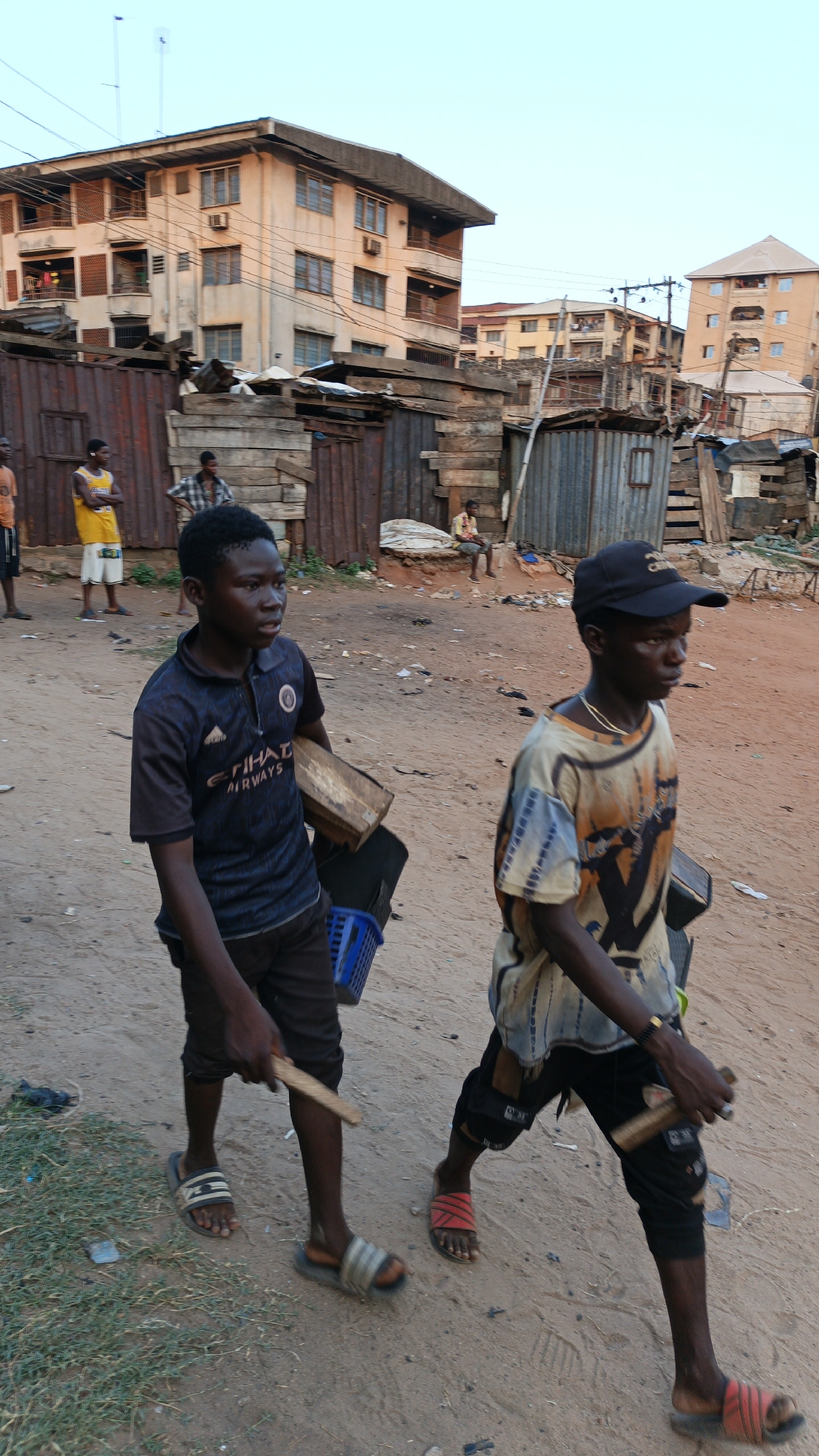 This vertical image captures a street scene in Onitsha, Nigeria, featuring two young males walking along a dirt road in the foreground. The boy on the left wears a dark blue polo shirt with "ETIHAD AIRWAYS" printed on it, dark shorts, and striped sandals. He carries a small wooden box, a blue plastic basket, and a wooden stick or paddle. Beside him, the boy on the right wears a black baseball cap, a patterned t-shirt with yellow, black, and white tones, dark pants, and red sandals. He holds two similar wooden sticks or paddles. Both boys appear focused as they walk.
The background is an urban landscape characterized by a mix of structures. Directly behind the boys are numerous makeshift stalls or dwellings constructed from corrugated metal sheets and wooden planks. Further back, multi-story concrete buildings with balconies and visible air conditioning units rise against a pale blue sky, suggesting late afternoon or early morning light. Several other individuals are visible standing or sitting among the background structures. The ground is unpaved dirt, with some patches of grass and scattered debris. The scene depicts daily life in a bustling, semi-urban environment.