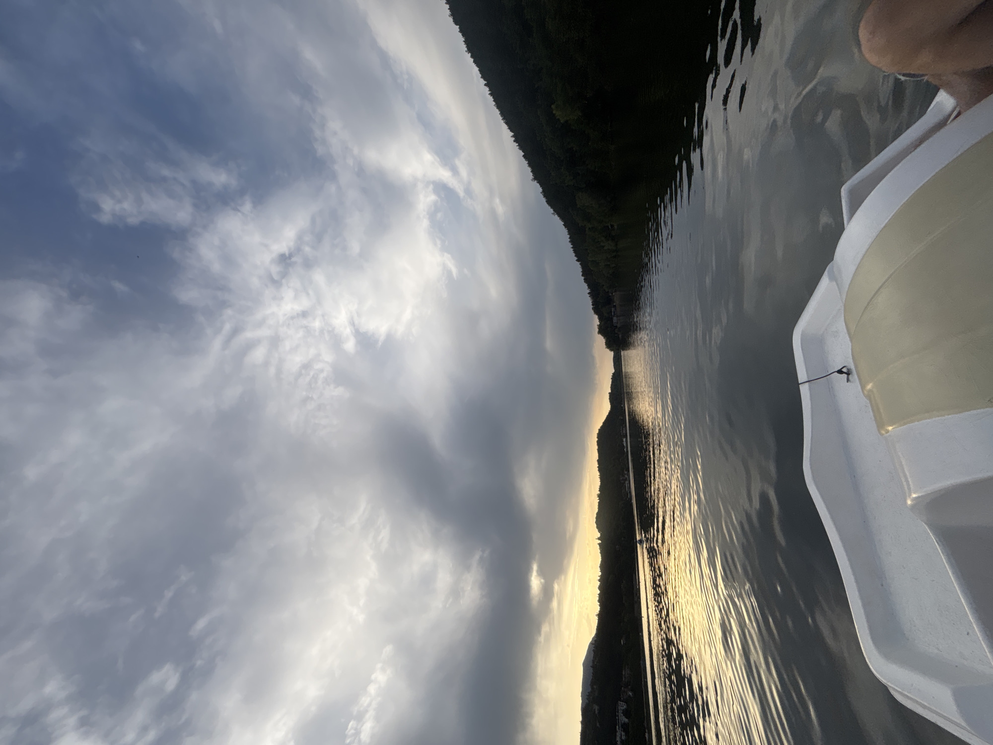 This is a first-person perspective shot taken from a pedal boat on the Pancharevo Lake in Pancharevo, Bulgaria. The sky is overcast with dramatic clouds, and the sun is setting, casting a golden glow on the water and the tree-covered hills in the distance. The water is calm, with gentle ripples reflecting the sky. The white pedal boat is in the foreground, and the legs of a person are visible on the right side of the frame. The scene evokes a sense of peacefulness and solitude.