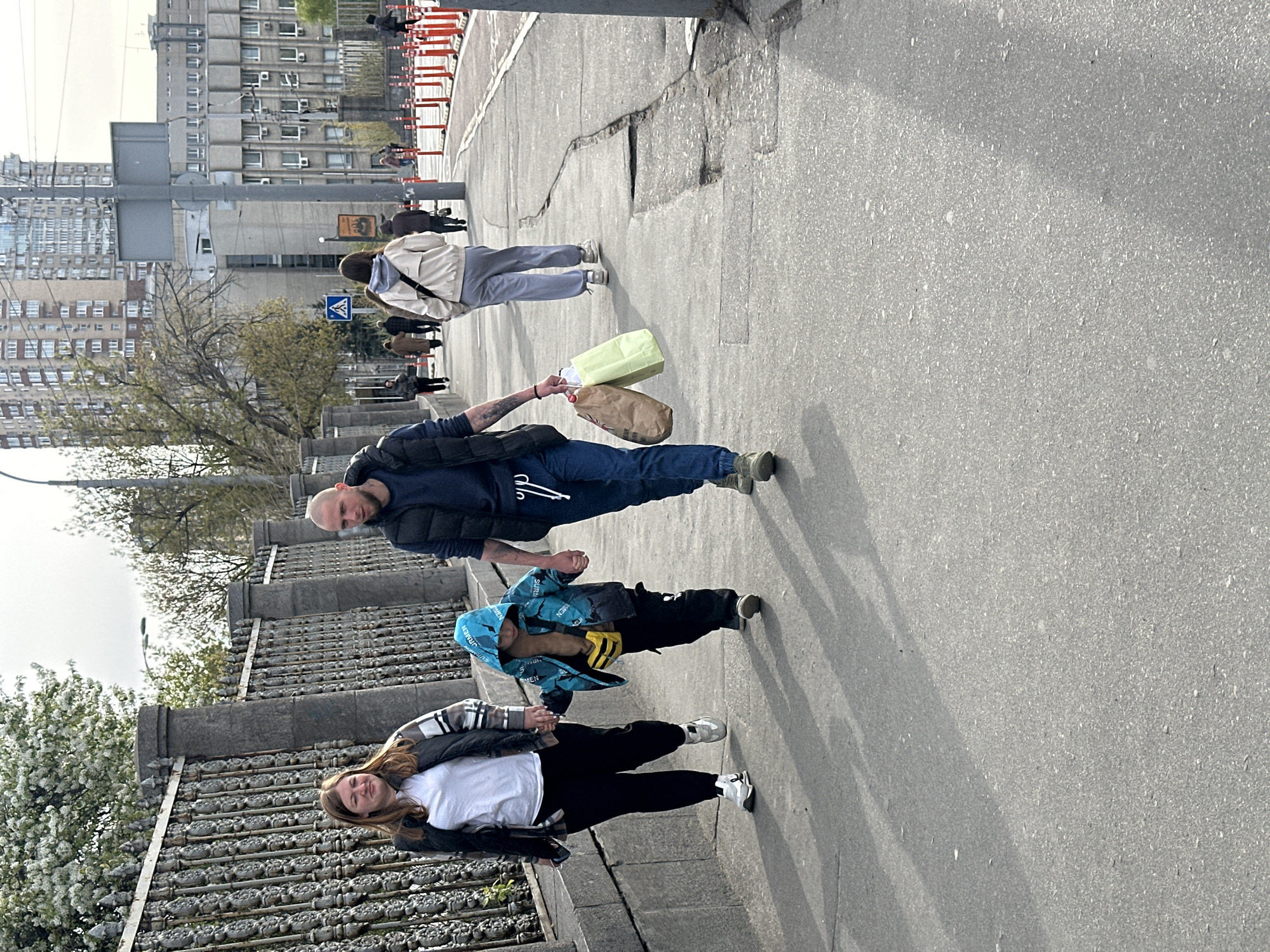 The image depicts a group of people walking along a sidewalk in Kyiv, Ukraine. The group consists of four individuals: a woman with long brown hair wearing a black and white jacket, a white t-shirt, and black pants; a child in a blue hooded jacket with a yellow fanny pack; a bald man in a navy blue puffy vest, a dark shirt, and jeans, holding two shopping bags; and a person further back, wearing a white jacket and gray pants. They appear to be walking along a bridge, indicated by the ornate metal railing. In the background, there are buildings, trees, and some traffic barricades. The sidewalk is cracked in places, suggesting wear and tear. Other pedestrians can be seen in the distance.