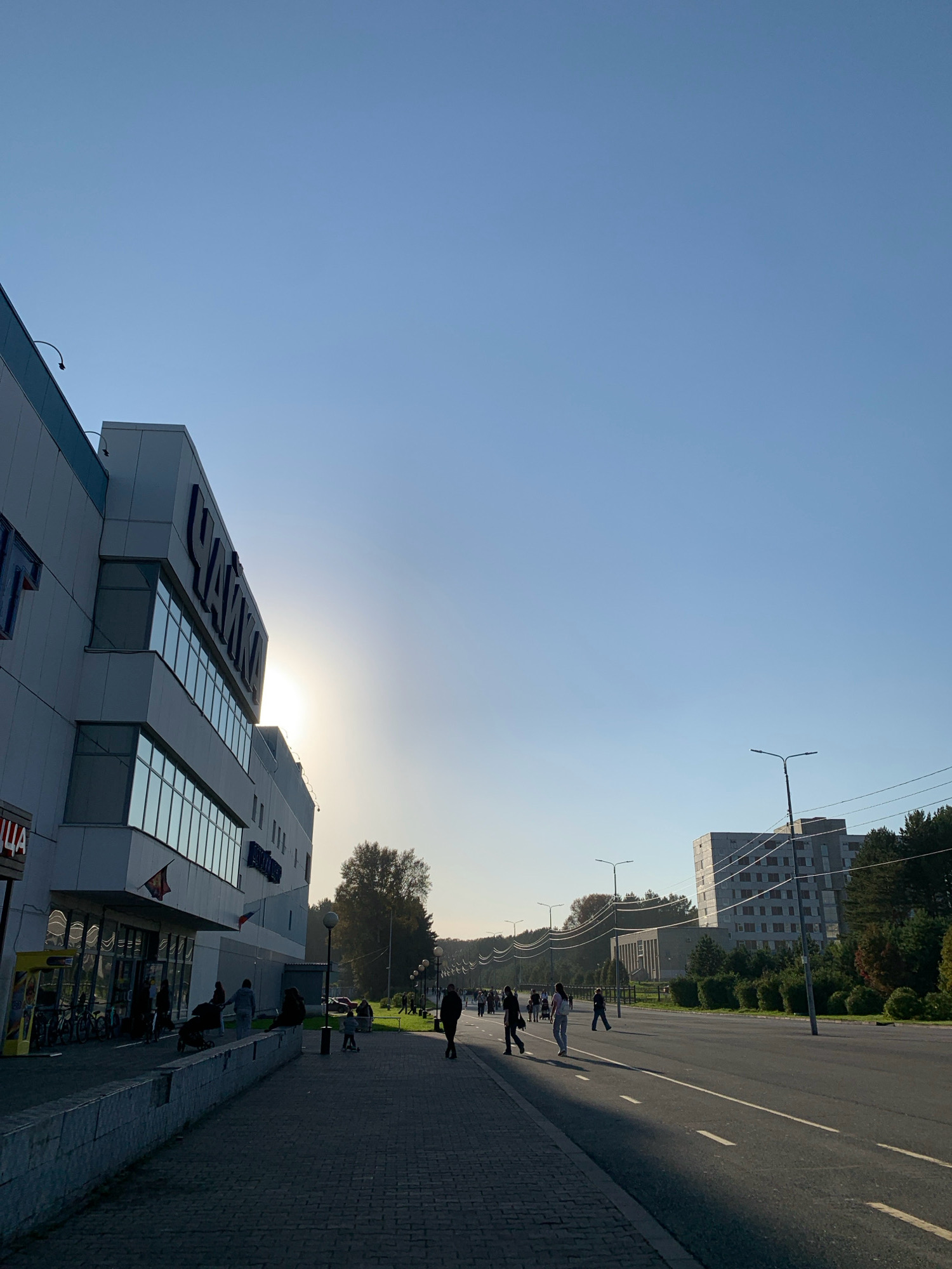 The image shows a wide street in a city, with a modern commercial building on the left and residential buildings in the distance. The sky is clear and blue. On the left is a large, light-colored building with several stories, featuring large windows and prominent signage that appears to read "ЧАЙКА" (CHAYKA). The facade of the building is partially in shadow, while the sun shines brightly behind it. A sidewalk runs along the front of the building, with several people walking or standing. Some are pushing strollers, suggesting families are present. Bicycles are parked outside the building, and there is a yellow vending kiosk. The street itself is paved and marked with white lines. Several streetlights are visible along the sidewalk and road. In the background, there are trees and more residential buildings, including a distinctive, modern-looking block of apartments with an unusual angular design. The scene appears to be set in the late afternoon or early evening, given the low angle of the sun and the shadows cast. The overall impression is one of a typical urban environment. The location is identified as Prokopyevsk, Russia, suggesting a view of a commercial and residential area within that city.