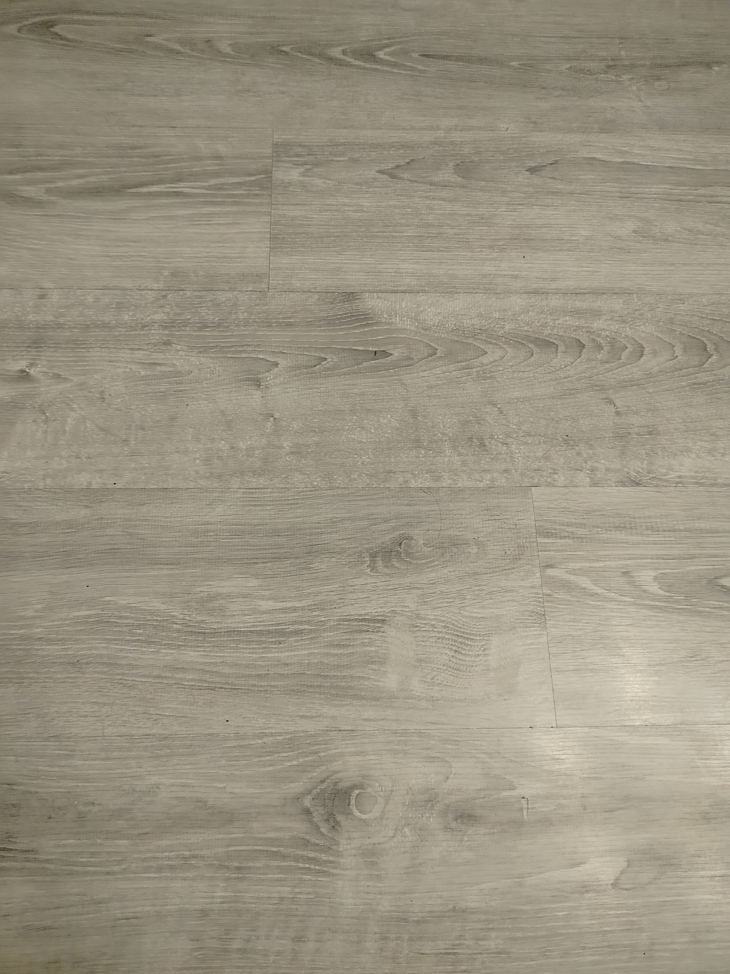 The image shows a close-up of a light grey wood-look floor. The planks run vertically in the image, with visible seams between the planks. The wood grain pattern is visible and includes knots and swirls. The scene does not contain information about Yerevan, Armenia, or any people, subjects, objects, actions, or interactions beyond the floor itself.
