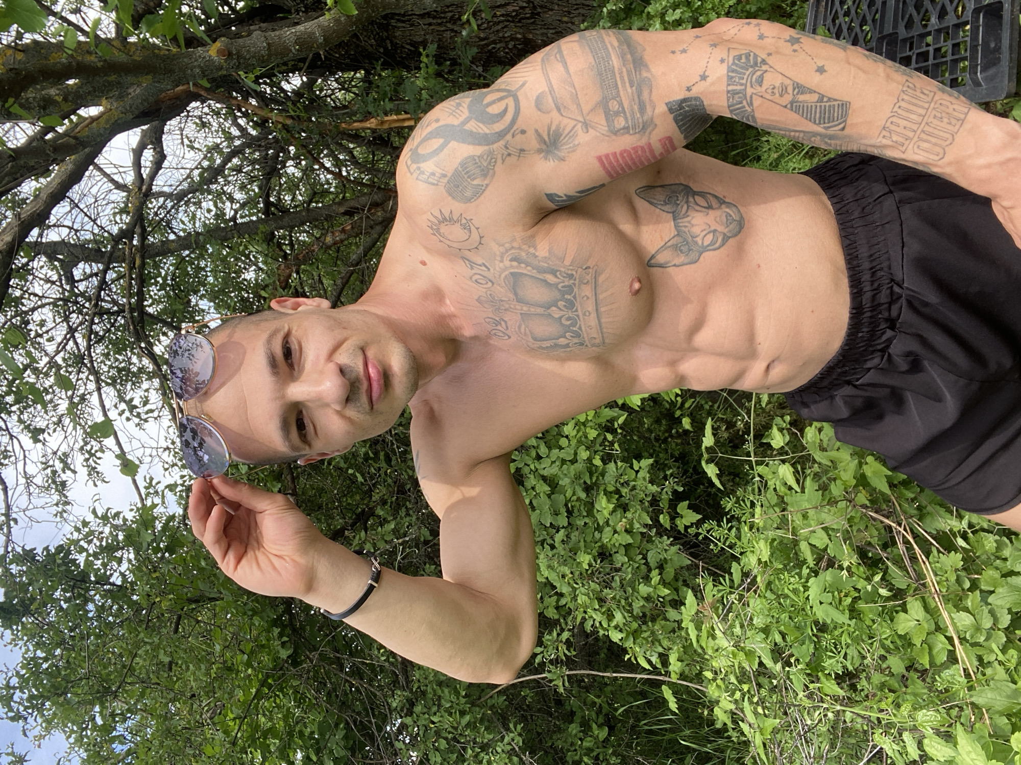 A shirtless man with tattoos on his torso and arms is standing in a grassy area near trees. He is wearing sunglasses, which he is holding in his left hand, and black shorts. The location is outdoors, with green foliage visible. The man is looking directly at the camera. It is daytime. There is no indication of the city of Pleven, Bulgaria, in the image.