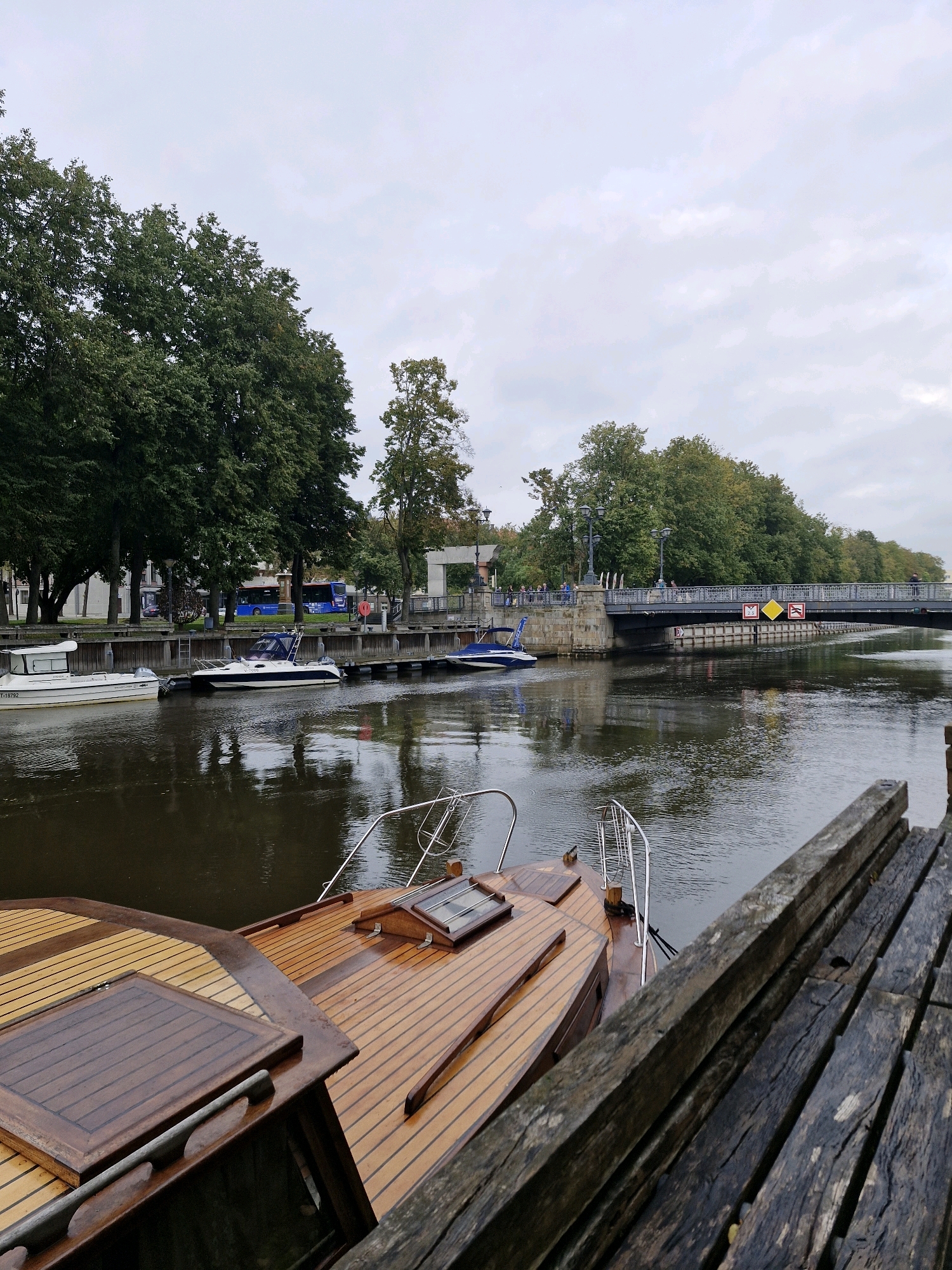 A view from a wooden dock shows several boats moored in a canal in Klaipėda, Lithuania. In the foreground, the stern of a wooden boat with a varnished deck is visible. Several other boats are docked further down the canal, including a white motorboat with a dark stripe and a blue and white motorboat. Across the canal, a treelined embankment is visible, with a pedestrian bridge spanning the water. People can be seen walking on the bridge. A blue bus and cars are visible on a road behind the trees on the embankment. The sky is overcast.