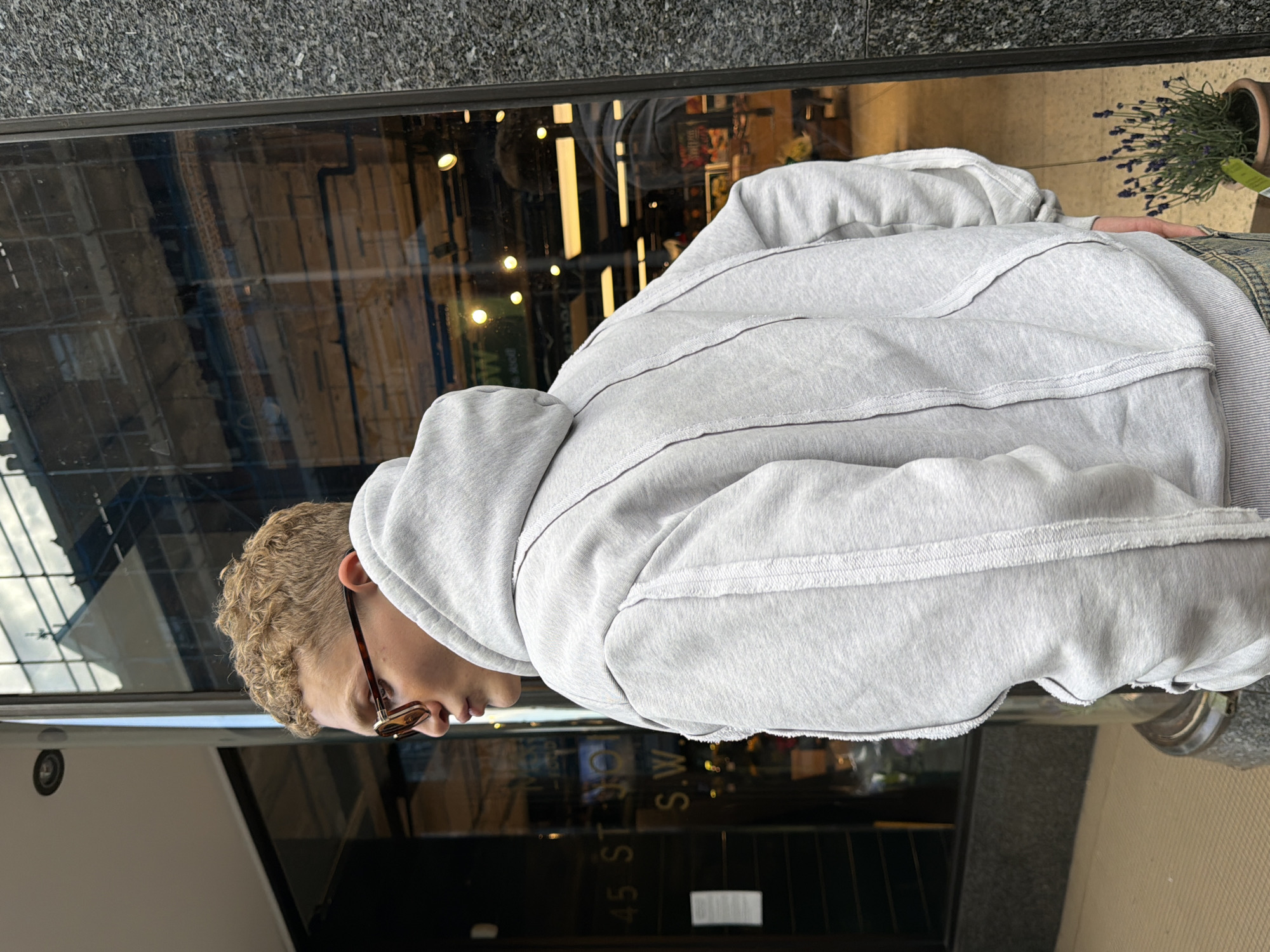 A person wearing a light grey hooded sweatshirt and sunglasses is standing outside a building with a reflective glass facade. The person is facing away from the camera, with their head turned to their left. The facade reflects the street, with shops and buildings visible. The person's hair is short and curly, and dyed a light blonde color. The person is wearing light-colored pants with a subtle pinstripe pattern. To the right of the person, a potted lavender plant is visible. The building has the numbers "45 ST" and "S.W." visible on the glass. The location is London, United Kingdom. The image is taken in an urban environment, with a grey stone wall on the right side of the frame. The lighting suggests it is daytime.