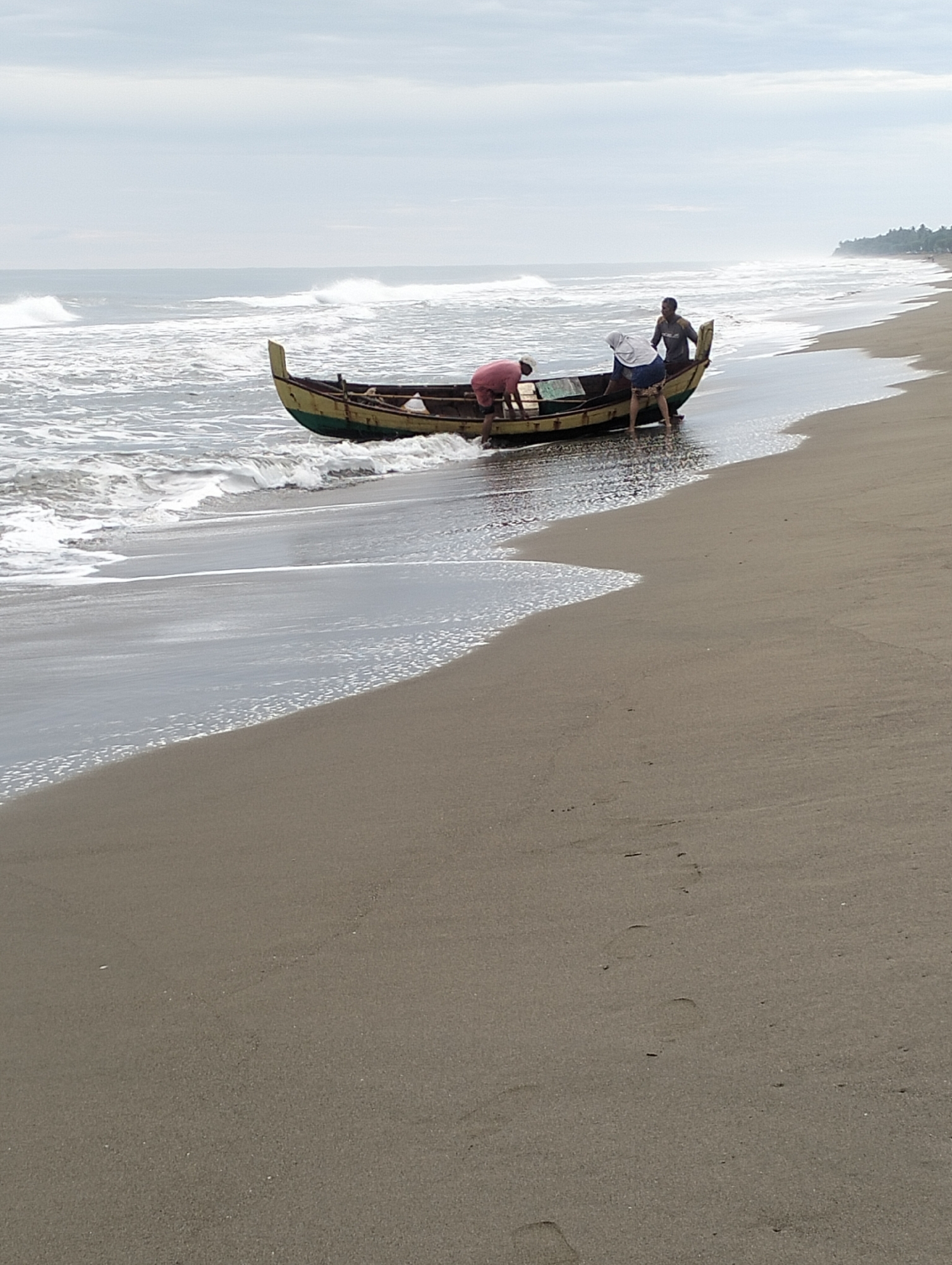 In Curehtunong, Indonesia, three individuals are engaged with a weathered, yellow and dark-colored wooden fishing boat partially submerged in shallow water on a sandy beach. The boat is oriented parallel to the shore, with its bow pointing towards the left and stern towards the right. One person, wearing a pink shirt and dark shorts, is positioned at the bow, leaning over the edge. Another person, in a light-colored shirt and dark shorts, is in the middle of the boat, also leaning. A third individual, in a grey shirt and dark shorts, stands at the stern, facing the boat. Gentle waves are lapping at the shore and the hull of the boat. The sky is overcast with grey clouds, and the sea stretches out to the horizon, with a tree line visible in the distance along the coast. The sand in the foreground is light brown and shows some scattered dark spots.