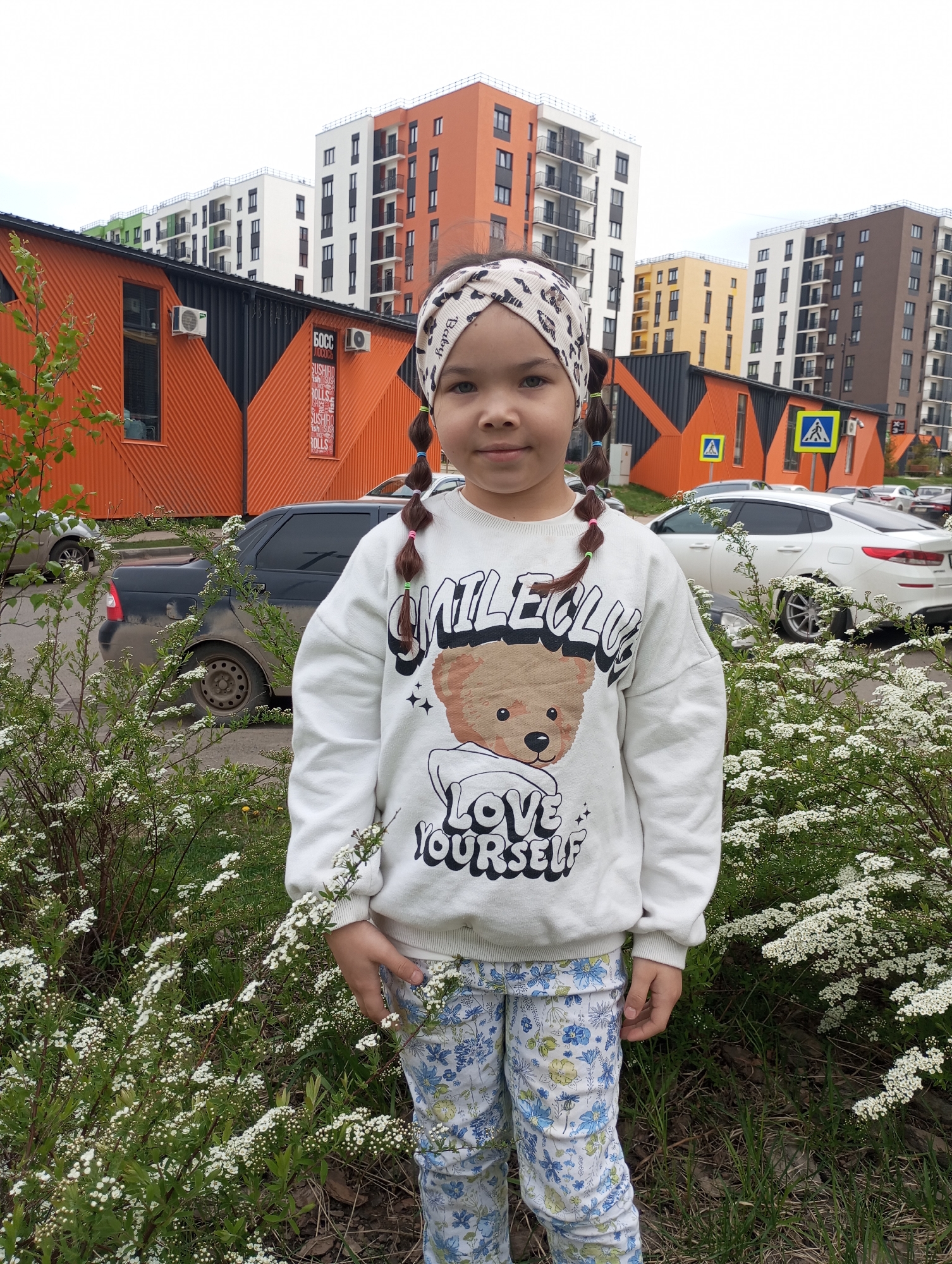 A young girl stands in a city setting. She has brown hair in pigtails with colored elastics. She wears a white sweatshirt with a bear graphic and the words "SMILECLUB LOVE YOURSELF" and blue patterned pants. She also wears a printed headband. Behind her, there are buildings with orange, black, white, green, yellow, and brown colors. Cars are parked in the background, and white flowering bushes are in the foreground. There is a crosswalk sign visible behind the girl. Based on the architectural style of the buildings, the location is likely Pervomaysky, Russia.