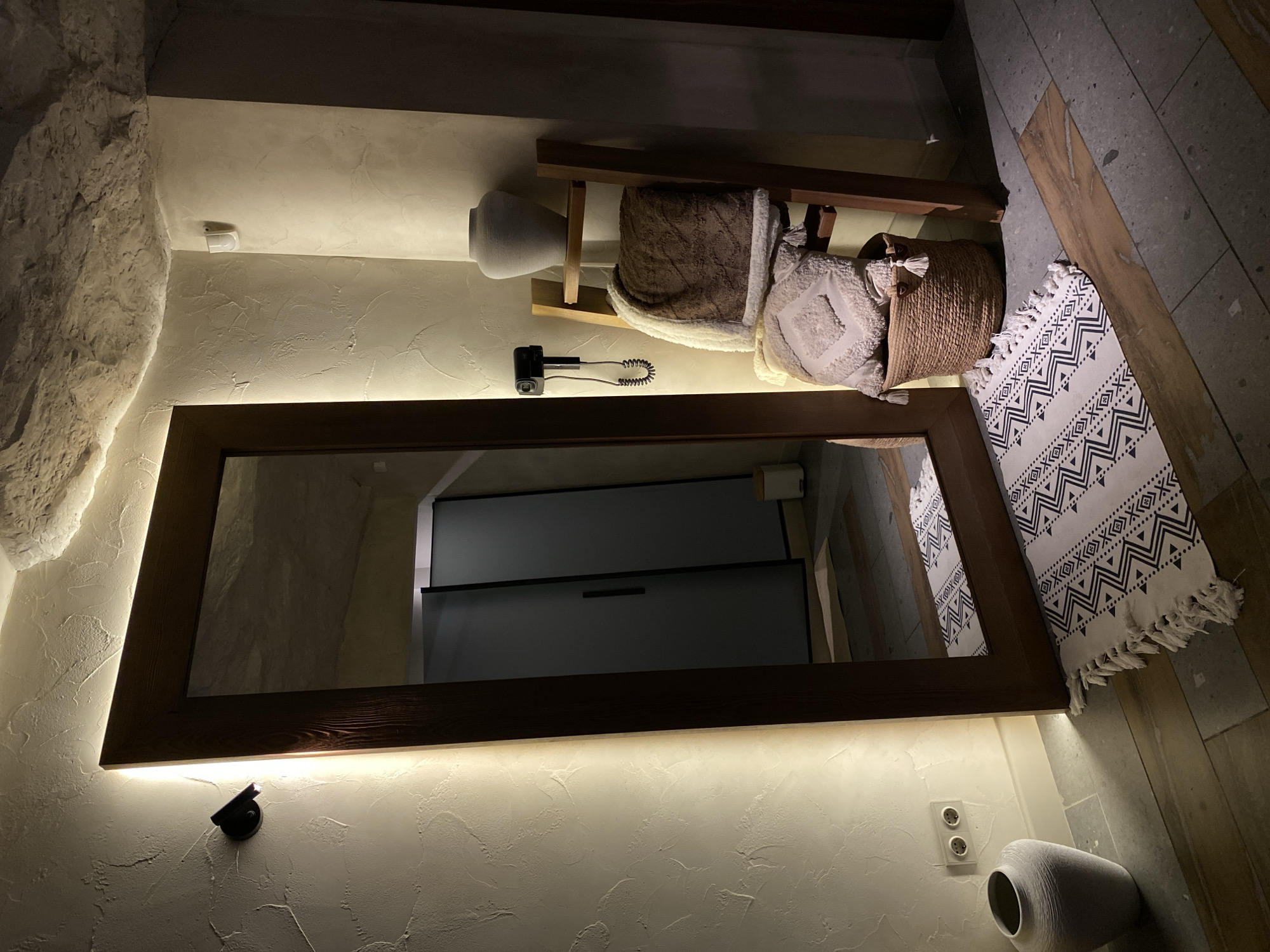 The image shows a bathroom interior with a large rectangular mirror dominating the scene. The mirror is framed in dark wood and backlit. Reflected in the mirror are gray cabinets. Above the mirror is a decorative arrangement consisting of a wooden shelf holding a light-colored vase, a brown rolled blanket, and a woven basket. The walls are textured in a light beige color. Below the mirror, near the floor, is a white vase. A black light fixture and an electrical outlet are visible on the wall. A patterned white and dark-colored rug is positioned near the right edge of the mirror. The floor appears to be tiled. There is no information to ascertain that it is in the city of Izhevsk, Russia.