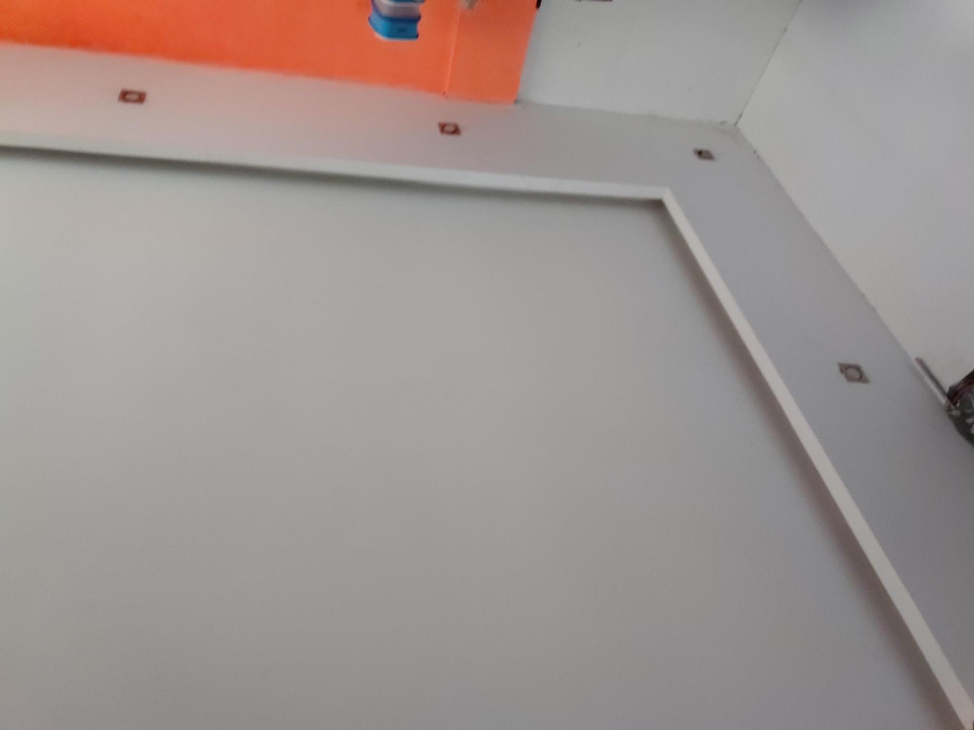 This interior shot captures an architectural detail of a room, looking upwards towards the ceiling and upper walls. The ceiling is a striking orange color, with a small blue object, possibly part of a light fixture or utility, hanging from it. Below the orange ceiling, a white border or soffit runs along the top of the walls. Embedded within this white border are several small, dark square openings, which appear to be recessed lighting fixtures. One such fixture is clearly visible in the upper left, and others are spaced out along the white trim.
The main focus of the image is a large, light grey rectangular panel on the wall, outlined by a clean white frame or trim. This panel dominates the central and lower portions of the frame. To the right of this panel, a white wall extends upwards, also featuring a recessed lighting fixture. In the far upper right corner, a darker, indistinct object, possibly tools or equipment, is partially visible.
The scene depicts a static interior space, possibly under construction or recently finished, given the clean and minimalist surfaces. There are no people or discernible activities taking place. No text is visible in the image. The overall aesthetic suggests a modern design with a bold use of color on the ceiling.