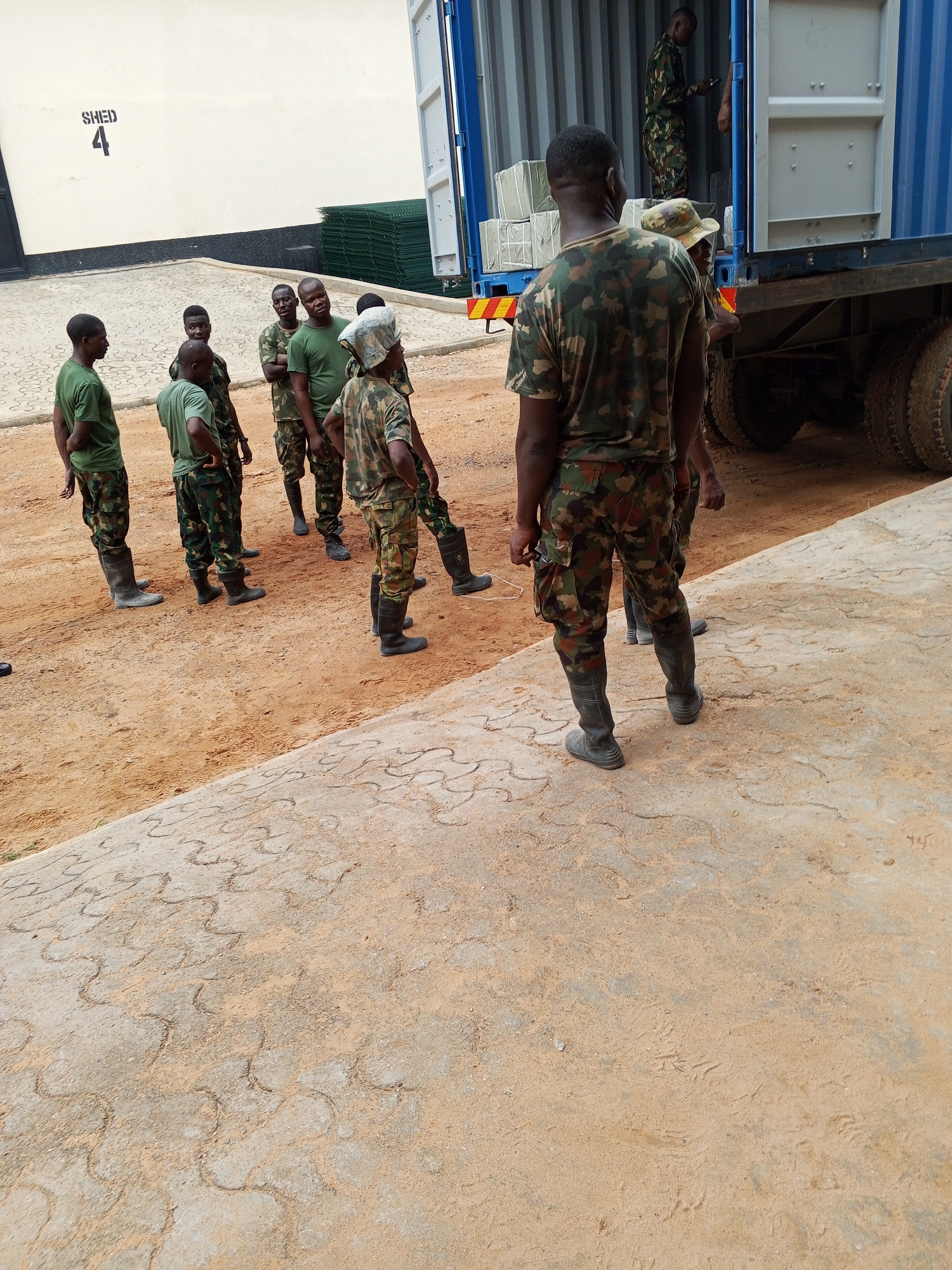 A group of soldiers are standing on a dirt and stone surface in front of a blue shipping container attached to a truck. Some of the soldiers are wearing camouflage uniforms and rubber boots, while others are in green shirts and camouflage pants. One soldier is standing in the shipping container. A building with "SHED 4" written on it is visible in the background. The location is Pasere, Nigeria.