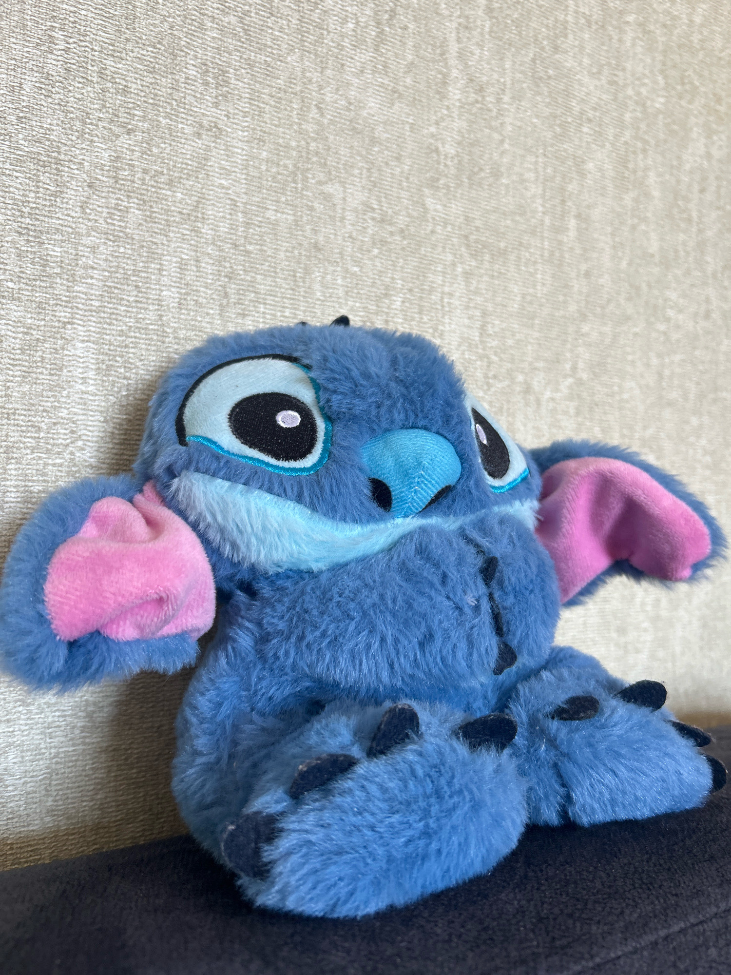 A plush toy resembling the character Stitch from Disney's Lilo & Stitch is positioned centrally in the frame, resting on a dark surface. The toy is predominantly blue with pink accents on its inner ears and paws. It has large black and white eyes with blue detailing around them and a blue nose. The background is a textured, light-colored wall. The image does not contain information to place it in Khyrdalan, Azerbaijan.