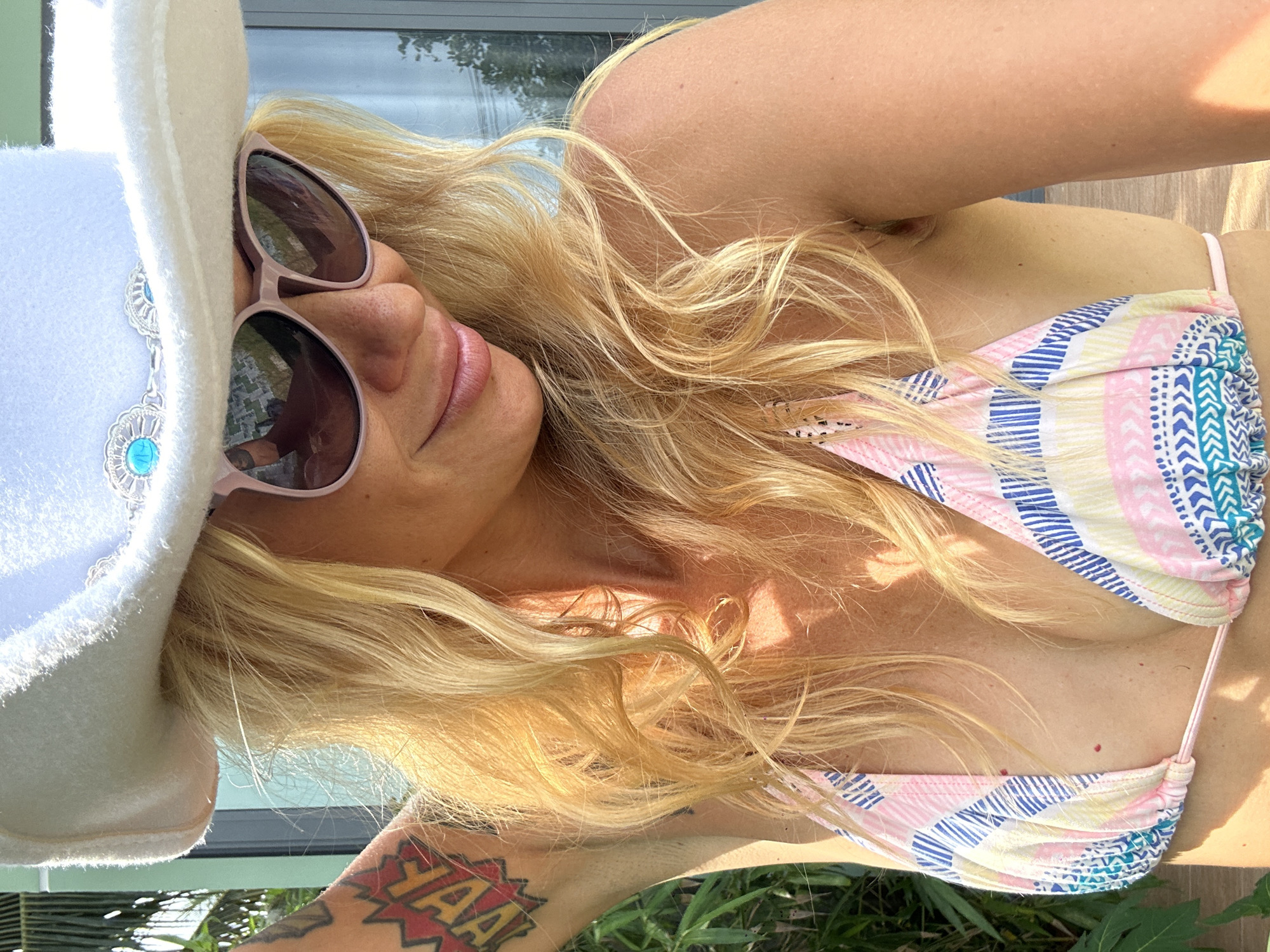 A woman with blonde hair and sunglasses is lying down outdoors, wearing a white cowboy hat and a colorful bikini. The sun is shining brightly, casting shadows and highlighting her skin. Her hair is loose and catches the light, and some of her chest and shoulder are visible. A tattoo is visible on her arm with the word "YAA!" in a comic-book style. The background suggests an outdoor setting with greenery and possibly a body of water or pool. The overall mood appears relaxed and summery, likely during daylight hours.