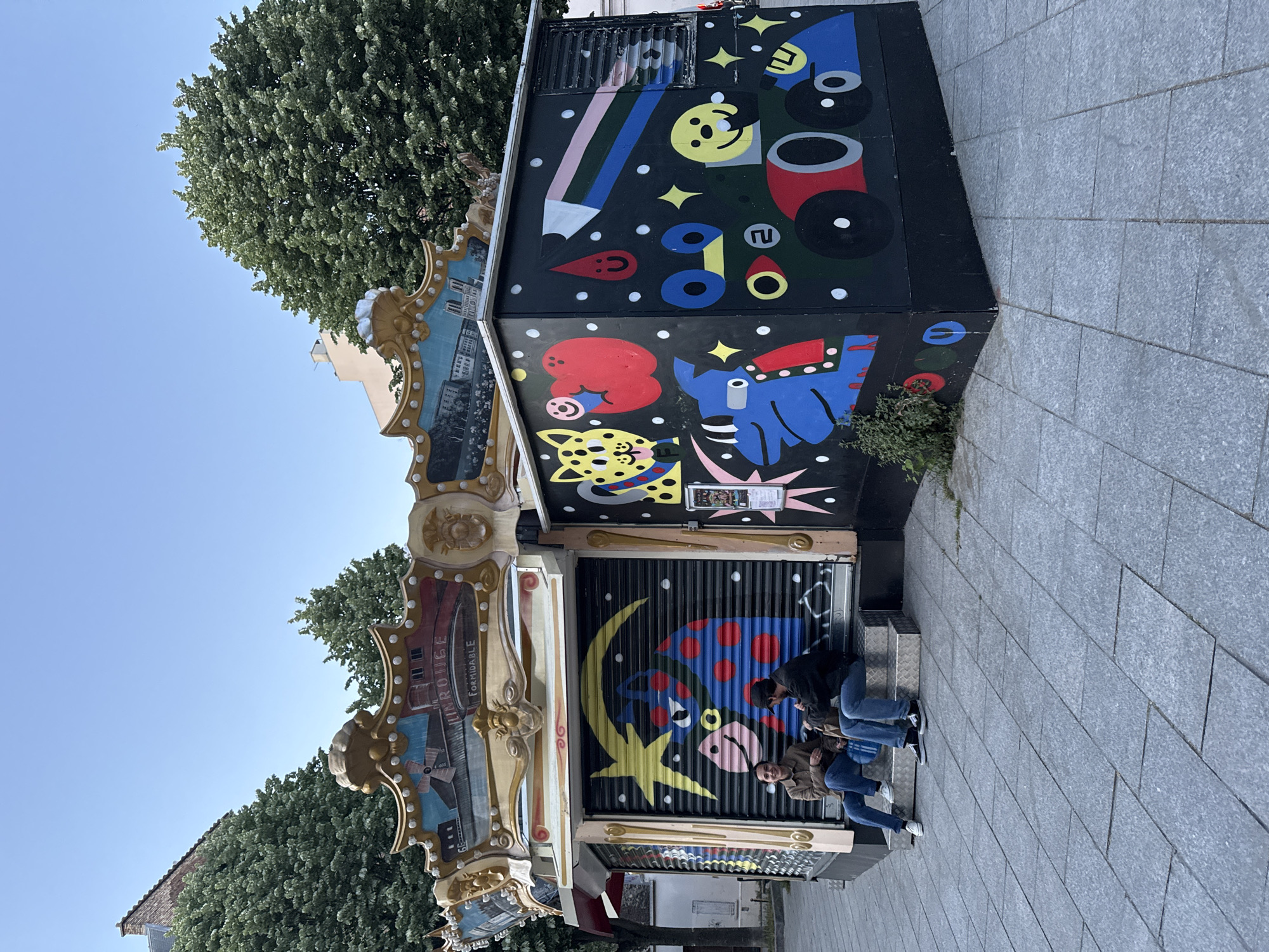 A vibrant daytime street scene in Paris, France, features a colorfully decorated kiosk or small building, set against a backdrop of lush green trees and a clear blue sky.
The kiosk's left section displays an ornate, gilded frame surrounding a painted cityscape, with visible French text "ROUGE" and "Formidable," suggesting a classic carousel or funfair aesthetic. The main facade of the kiosk is dark, adorned with playful, abstract graffiti or murals. These include a large pencil, a yellow smiley face, red and blue circular elements, a red blob with a face, a blue elephant-like creature, and a yellow leopard-like face, all interspersed with stars and white dots. Below this, a corrugated metal shutter, presumably closed, is painted with a large yellow crescent moon and a blue, red-spotted creature that resembles a ladybug.
Two young men are casually seated on steps or a low ledge in front of the closed shutter. They are dressed in everyday clothing, including jeans and jackets, with one looking towards the camera. The ground in the foreground is a paved sidewalk of light grey square tiles. In the background, dense green foliage of trees (likely lime or linden) extends towards the top of the frame, underneath a bright blue sky. A light-colored building is partially visible behind the trees. The overall impression is one of urban vibrancy and relaxed atmosphere on a sunny day.