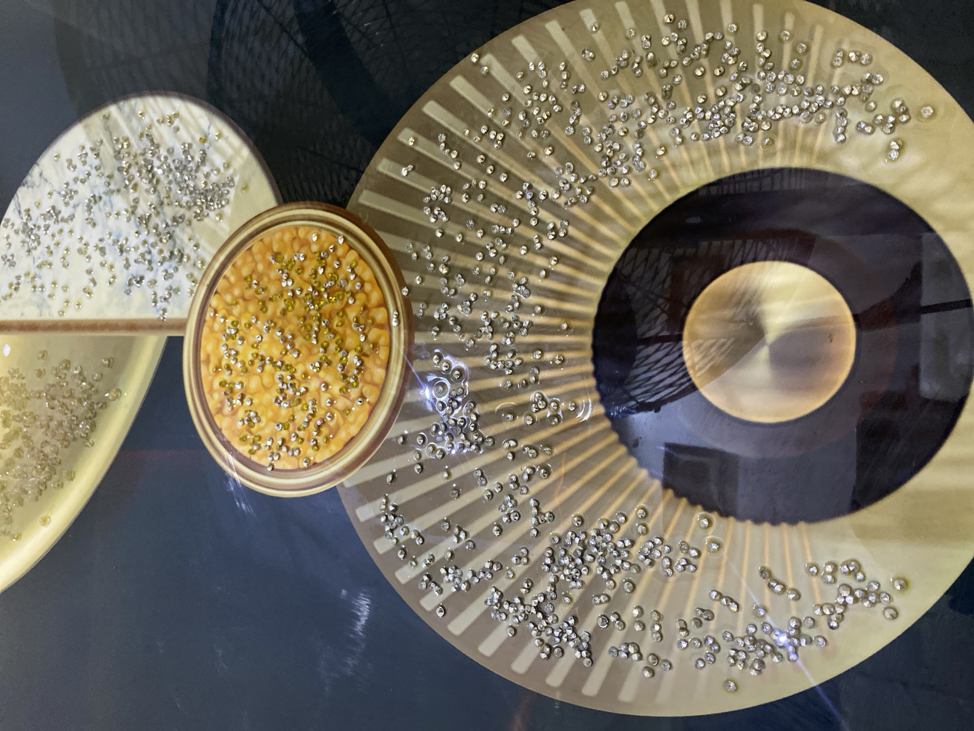 The image is a close-up, top-down view of an abstract artwork. The artwork features several overlapping circular and fan-shaped elements, primarily in shades of gold and brown. These elements are adorned with numerous small, sparkling rhinestones or crystals, scattered across their surfaces. One prominent circular element in the upper center appears to contain a pattern resembling corn kernels, also embellished with similar stones. The background is a dark, possibly black, surface. There is no indication of people, actions, or interactions within the image, nor any recognizable landmarks or environmental context that would suggest a location in Giginyu, Nigeria.
