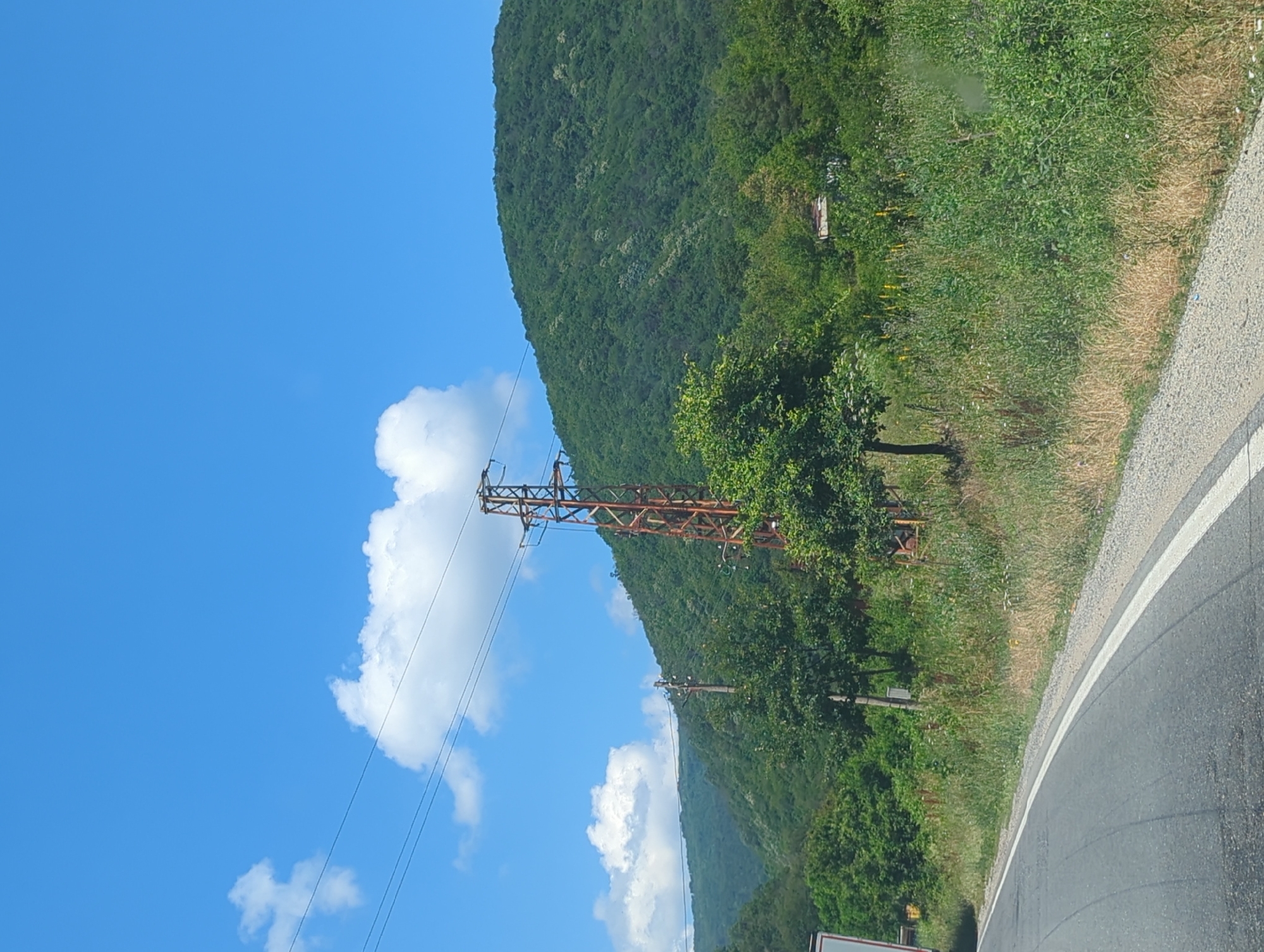 The image depicts a rural roadside scene in Pchelinovo, Bulgaria, under a clear blue sky with scattered white clouds. A paved road with a white line marking curves through the foreground. To the right of the road, a steep, verdant hillside is covered in dense green trees and foliage. A tall, rusted metal power pylon stands prominently on the hillside, with electrical wires extending across the frame. In the mid-ground, various trees and vegetation line the roadside, with a small, rustic building partially visible amidst the greenery. The overall impression is one of a scenic, undeveloped natural landscape with utilitarian infrastructure.