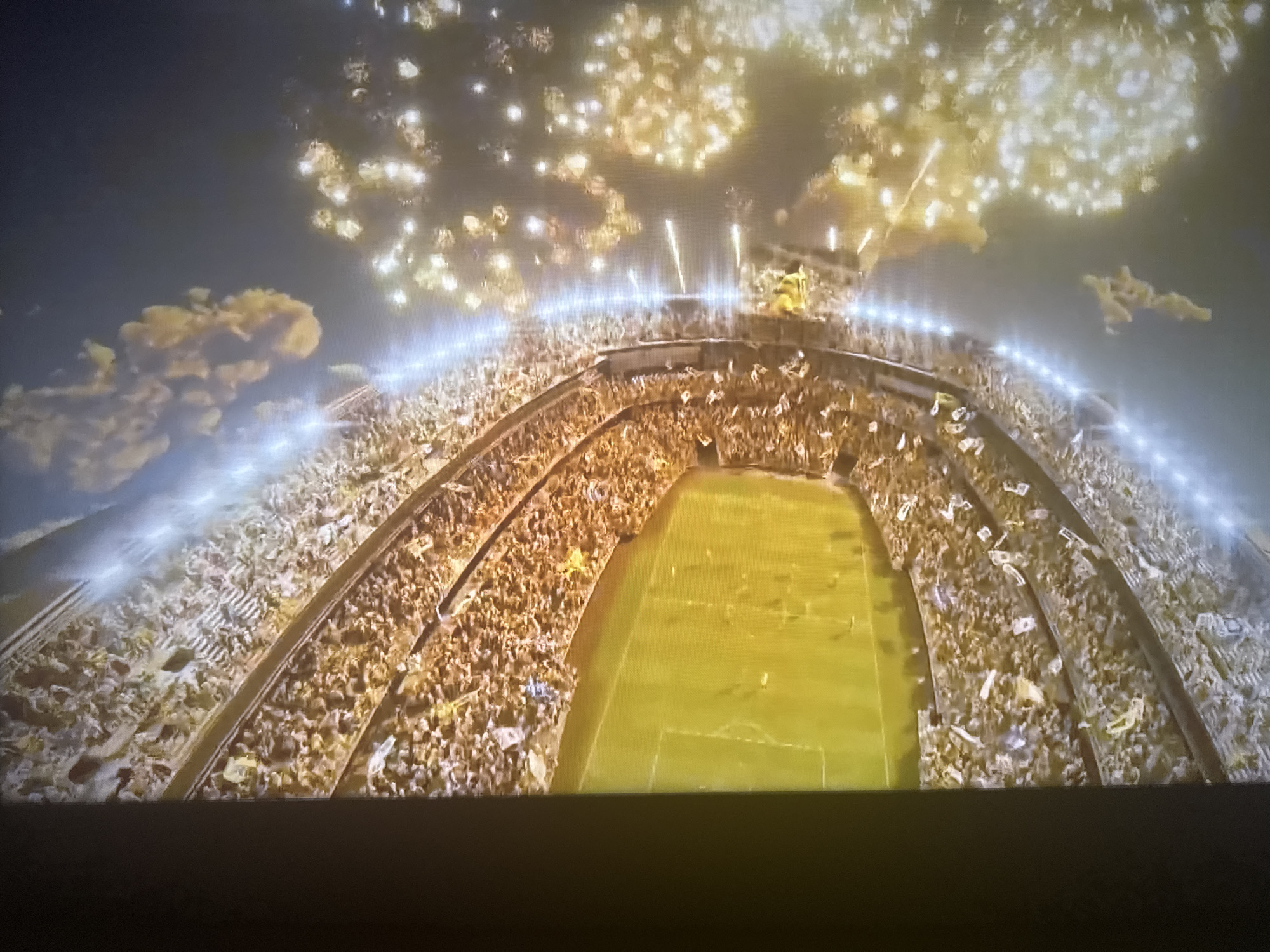 An aerial shot of a brightly lit football stadium at night, packed with a roaring crowd. Fireworks explode in the dark sky above, casting a warm, golden glow over the scene. The stadium's green turf is clearly visible, with players in yellow uniforms visible on the field. The stands are filled with spectators, many of whom are waving white flags, creating a celebratory atmosphere. The stadium lights create a bright, illuminated line around the top of the stands. The overall impression is one of excitement and festivity.