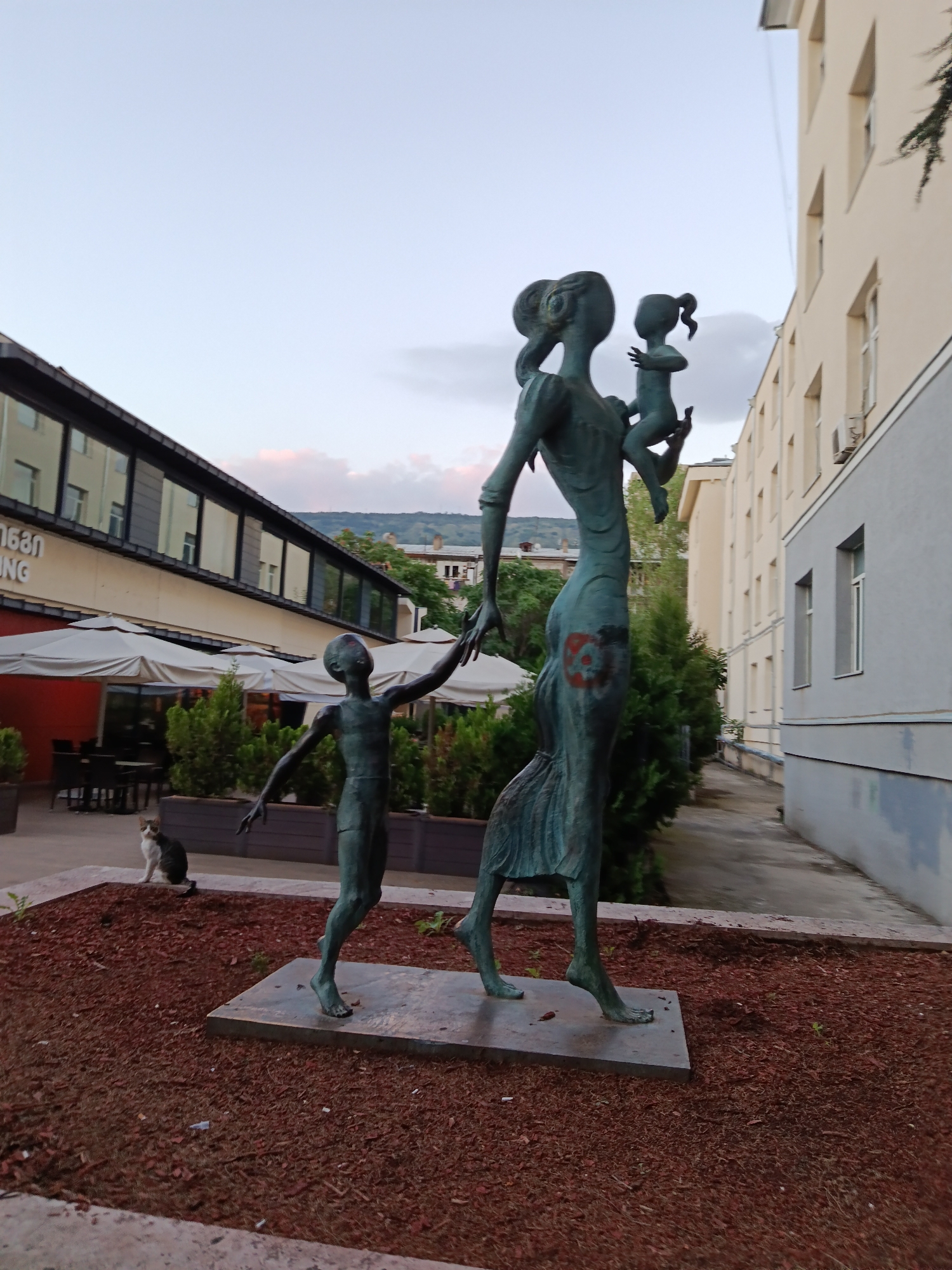 A full-shot, eye-level outdoor photograph depicts a bronze statue of a woman holding a baby and walking with a young boy. The statue is situated on a stone pedestal surrounded by red mulch and scattered green sprouts. The woman, in the foreground, faces right and is depicted with exaggerated features and a flowing dress. She holds a baby in her left arm. To her left, a boy with outstretched arms walks beside her. In the background, a multi-story building with cream-colored walls and numerous windows stands to the right. To the left of the statue, another building with a dark facade and large windows is visible. Several white umbrellas are set up in front of the building. A calico cat is perched on the left edge of the mulch, looking towards the statues. The sky above is a pale blue with faint clouds.