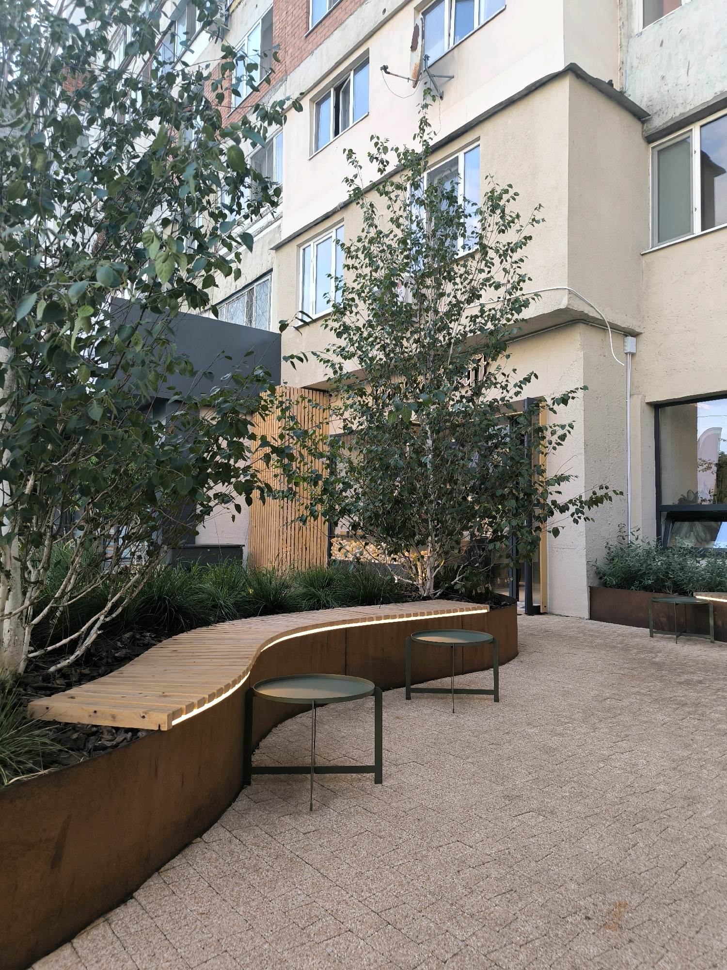 The image shows an outdoor seating area in front of a multi-story building. A curved wooden bench with integrated lighting is the focal point of the foreground, bordered by a Corten steel planter. Two small, green metal tables are placed on a light-colored, textured pavement. Behind the bench, there are trees with thin, pale trunks and lush green foliage, along with dense green ground cover. A wooden slatted wall forms a backdrop in the mid-ground, behind which a dark-colored structure is visible. The building itself appears to be residential, with a mix of brick and light-colored plastered facades and numerous windows. The scene is captured from a low angle, emphasizing the seating area and the surrounding landscaping. The overall impression is that of a modern, designed urban space intended for relaxation.