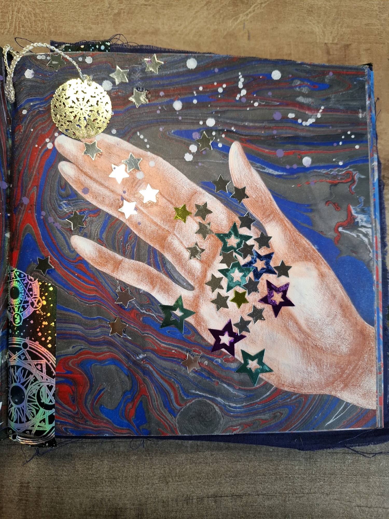 The image is a close-up, top-down view of an open art journal spread on a wooden surface. The journal page features a paint pour background with swirling patterns of dark gray, red, and blue. A painted rendering of an open hand, with skin tones ranging from peach to light brown, is placed centrally on the page, palm facing upwards. Scattered across the hand and the background are numerous small, metallic star-shaped sequins in various colors, including silver, gold, green, teal, and purple. A small, intricately patterned gold ornament, resembling a filigree ball, hangs from a silver string above the hand. To the left of the hand, a vertical strip of holographic material with circular and geometric patterns is adhered to the journal's edge. The journal is bound with dark blue fabric, visible at the bottom and left edges. The overall impression is one of artistic expression with celestial and mystical themes. The location in the city of Tyumen, Russia, is not discernible from the image.