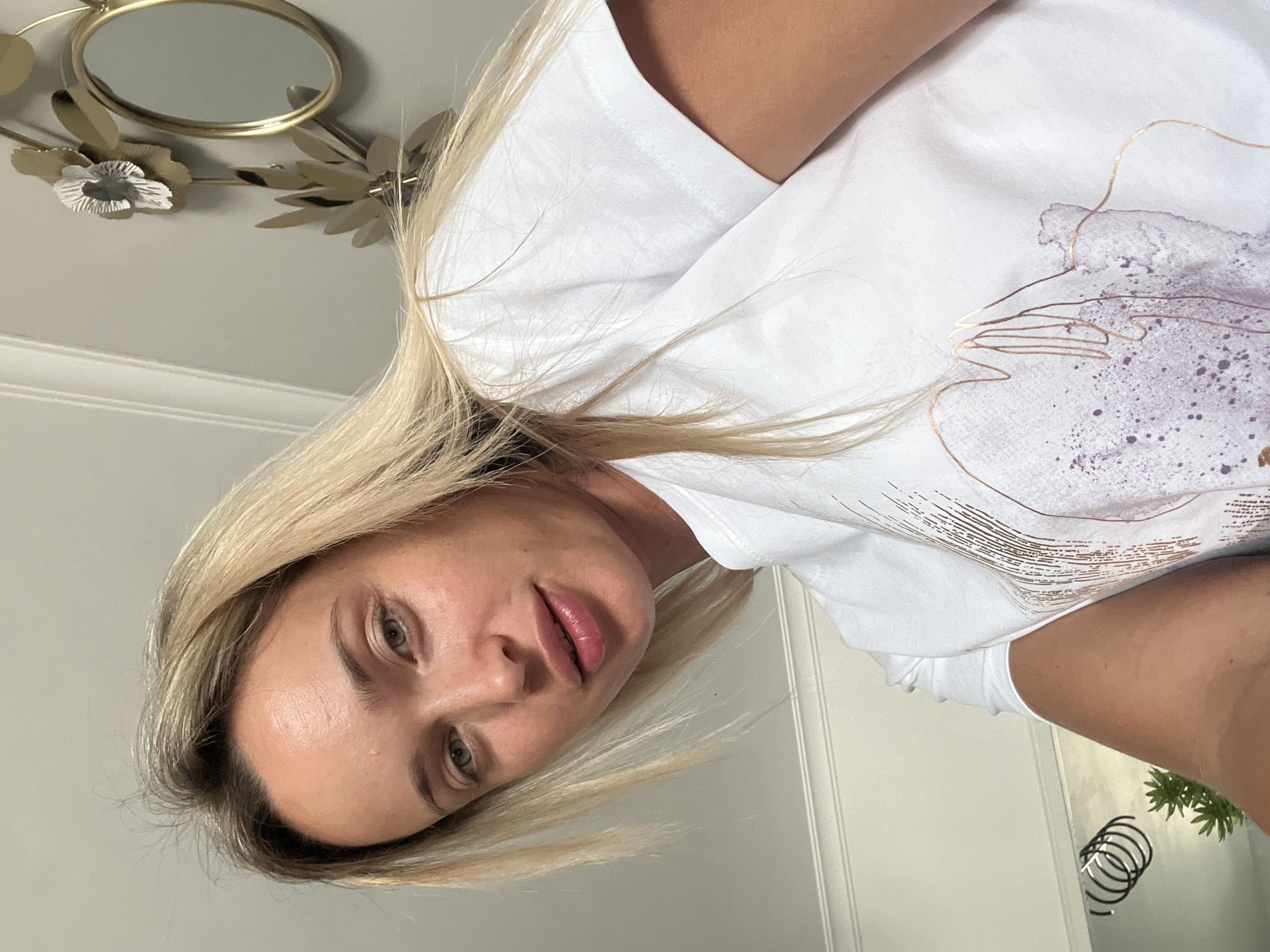 A close-up, selfie-style shot shows a woman with blonde hair and light skin wearing a white t-shirt. Her face is angled towards the camera, and she has light green eyes and pink lips. The background is a plain, light-colored wall with a gold decorative wall piece featuring a circular mirror and floral accents to the right of the woman. To the left, a white molding runs along the wall. In the lower left corner, a reflection in a mirror shows a black coiled object and a small green plant. The lighting is soft, suggesting an indoor environment. There is no indication of the specific location within Sharm El Sheikh, Egypt.
