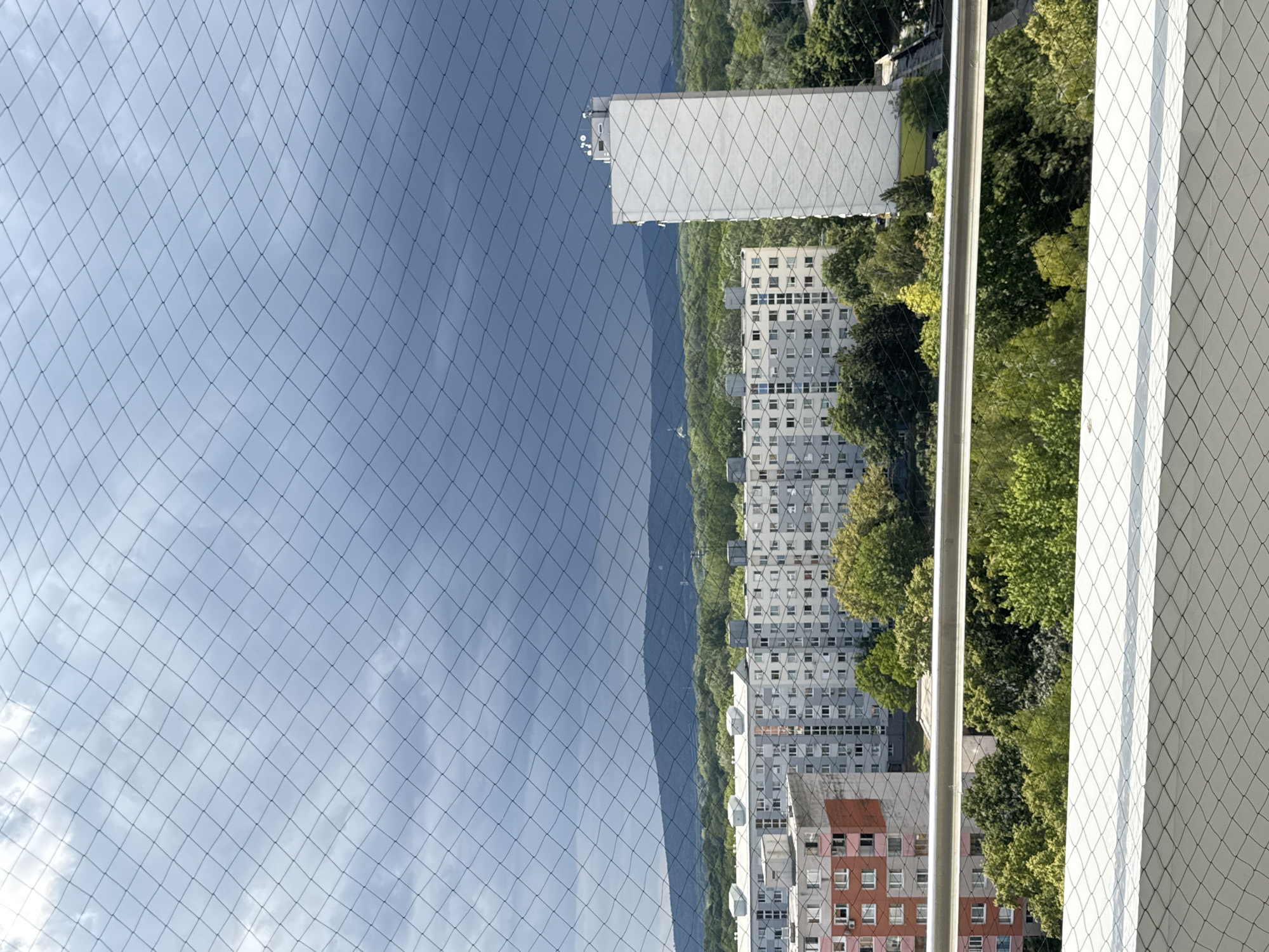 The image presents a view of a city landscape in Bratislava, Slovakia, seen through a fine mesh netting, likely a balcony safety net. The foreground is dominated by the white netting and a portion of a railing. Beyond this, a dense canopy of green trees occupies the mid-ground.
Behind the trees, several apartment buildings are visible. The most prominent is a long, grey building with numerous windows, extending across the frame. To its right, a tall, slender white building stands out, also with a pattern of windows. Another, shorter apartment building with orange and grey sections is visible to the left of the grey building.
In the background, rolling hills covered in green foliage form a distinct mountain range, under a dramatic sky. The sky is a mix of dark, overcast clouds and lighter, brighter patches, suggesting approaching or receding weather. The overall impression is that of an urban environment nestled amongst nature, viewed from a height.