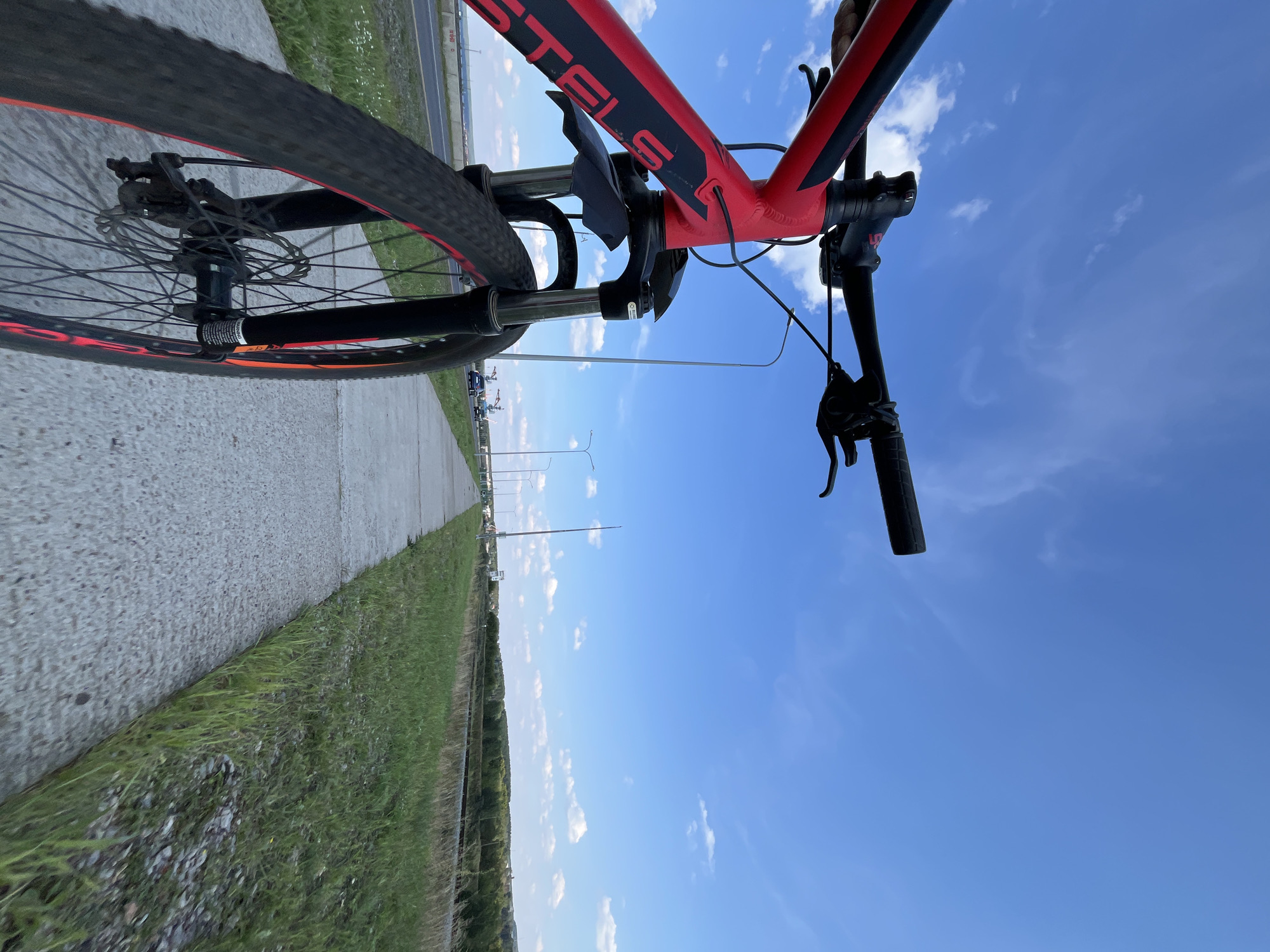 The image shows a red and black bicycle with a wide-angle perspective, positioned on a paved pathway next to a road. The bicycle's front wheel and handlebars are in focus, with the brand "STELS" visible on the frame. The pathway runs parallel to a road with a few cars visible in the distance. Beyond the road, there is a strip of green grass and trees, leading to a distant treeline under a bright blue sky with scattered white clouds. The location is the city of Chaikovsky, Russia, specifically along the pathway adjacent to the road and a grassy area. There are no people visible in the image. The scene appears to be outdoors during the daytime.