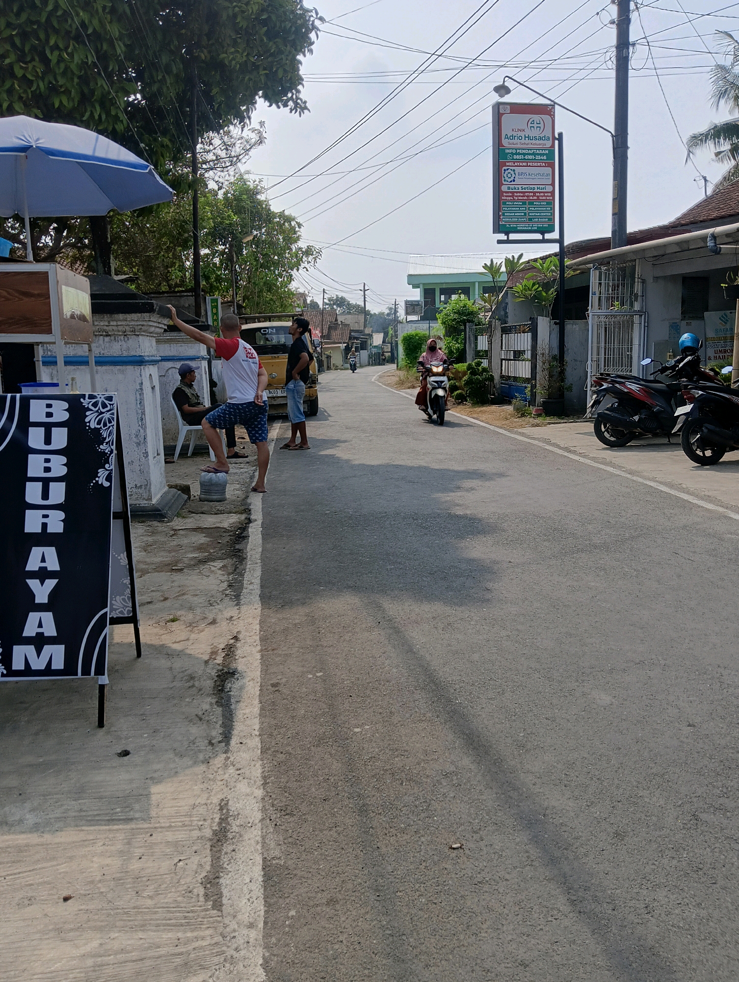 The image depicts a street scene in Sokaraja, Indonesia, under a clear sky during daylight. In the foreground on the left, a black A-frame sign displays the word "BUBURAYAM" in large white capital letters, indicating a food establishment. A blue and white umbrella is positioned behind the sign.
Several individuals are present on the side of the road. A man in a red and white shirt and patterned shorts is standing on a block, with his right arm extended upwards as if taking a selfie or pointing. Another man in a black t-shirt and blue shorts stands nearby, looking towards the street. A third person is seated on a white chair further back.
A yellow truck is parked on the side of the road. In the middle ground, a woman wearing a pink headscarf is riding a white scooter down the street. Further down the road, two motorcycles are parked on the right side.
A prominent sign for "KLINIK ADRIQ HUSADA" (Adria Husada Clinic) is visible on a pole, displaying information about the clinic's services, operating hours, and insurance coverage. The surrounding environment includes residential buildings and vegetation. The road surface is asphalt.