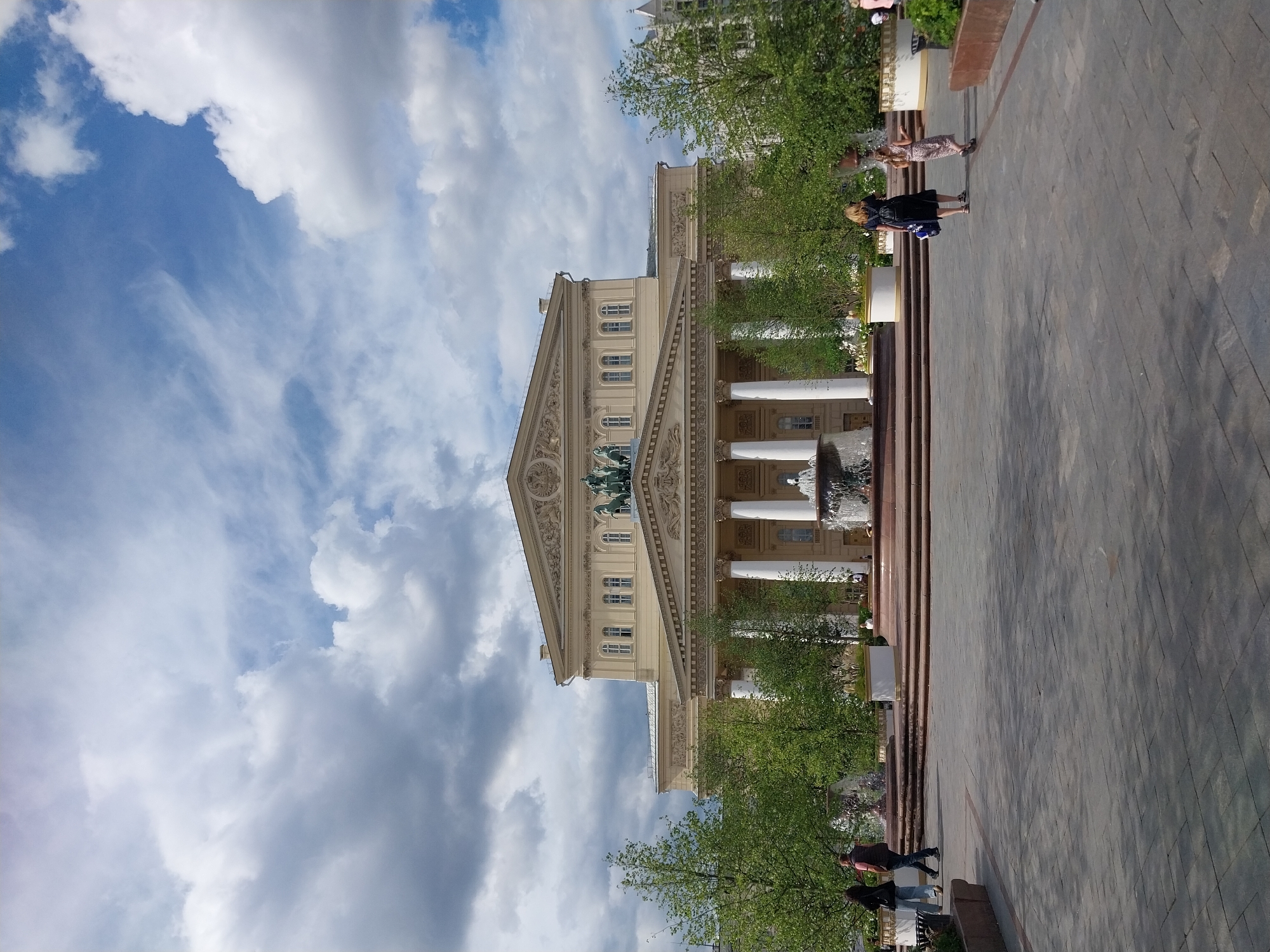 The image shows the Bolshoi Theatre in Moscow, Russia. The building is a grand neoclassical structure with a triangular pediment adorned with sculptures. White columns support the portico, and the facade is decorated with ornate carvings. The sky above is a mix of blue and white clouds, suggesting a partly cloudy day. Lush green trees surround the building, adding to the picturesque scene.
In the foreground, a paved plaza stretches out, with people casually strolling and observing the architecture. A fountain is visible, its water adding a dynamic element to the scene. The time of day appears to be afternoon, indicated by the lighting. The overall atmosphere is peaceful and inviting, capturing the beauty of this iconic landmark in Russia's capital. The people in the image are dressed in casual attire, suggesting a typical day for tourists and locals alike. The architecture and the presence of trees and a fountain indicate an urban park or public square setting. There is no visible text in the image.