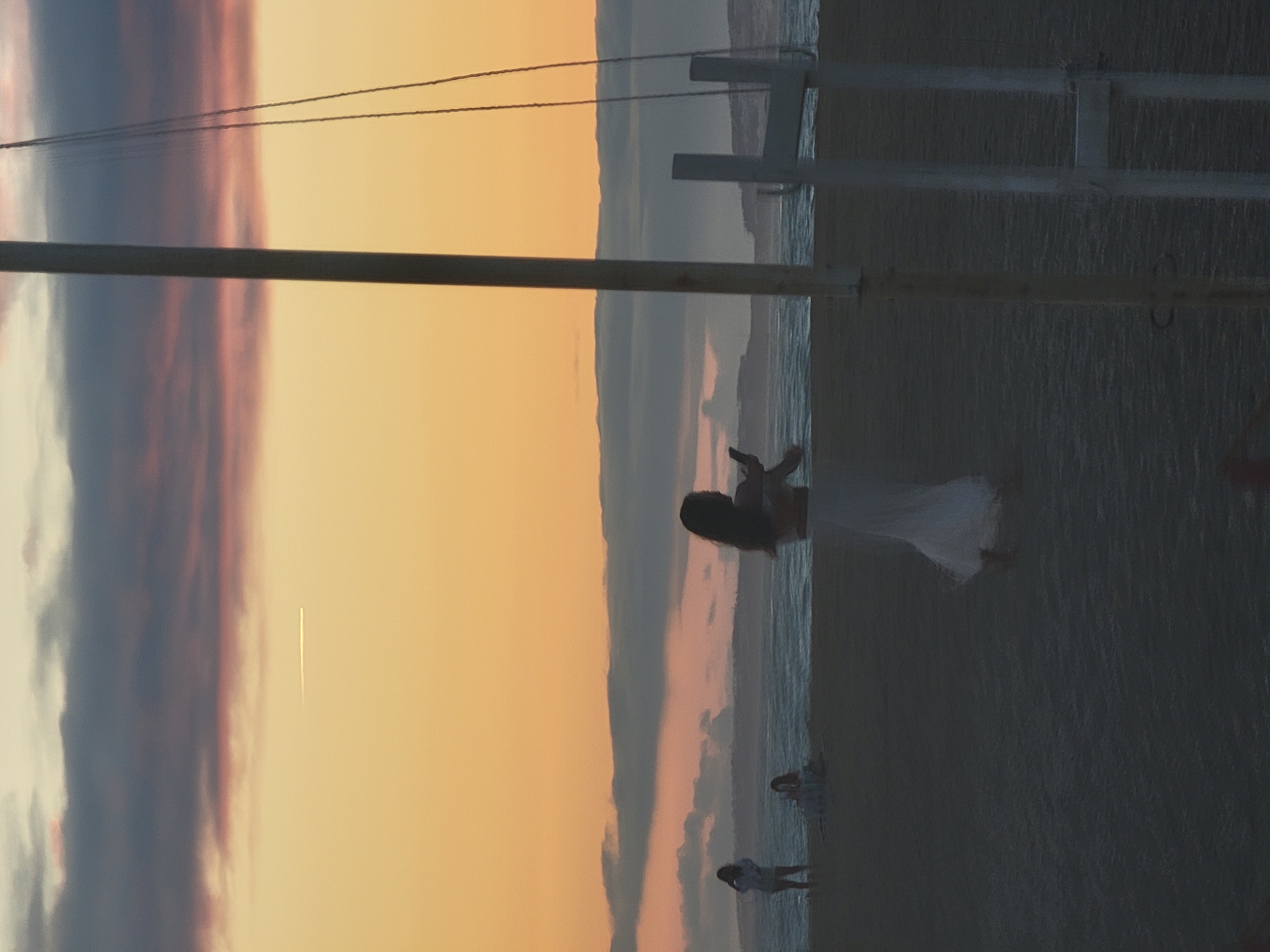 The image depicts a beach scene at sunset in Quarteira, Portugal. In the foreground, a person is silhouetted against the sky, holding a phone. To the left, two other figures are visible on the beach. The sky is a gradient of orange and pink, with clouds and a faint contrail from an aircraft. The ocean is in the background, with waves breaking on the shore. A tall pole, likely part of a beach structure, stands in the mid-ground, with ropes and part of a frame visible to its right. The sand is dark and textured, suggesting it is wet or damp.