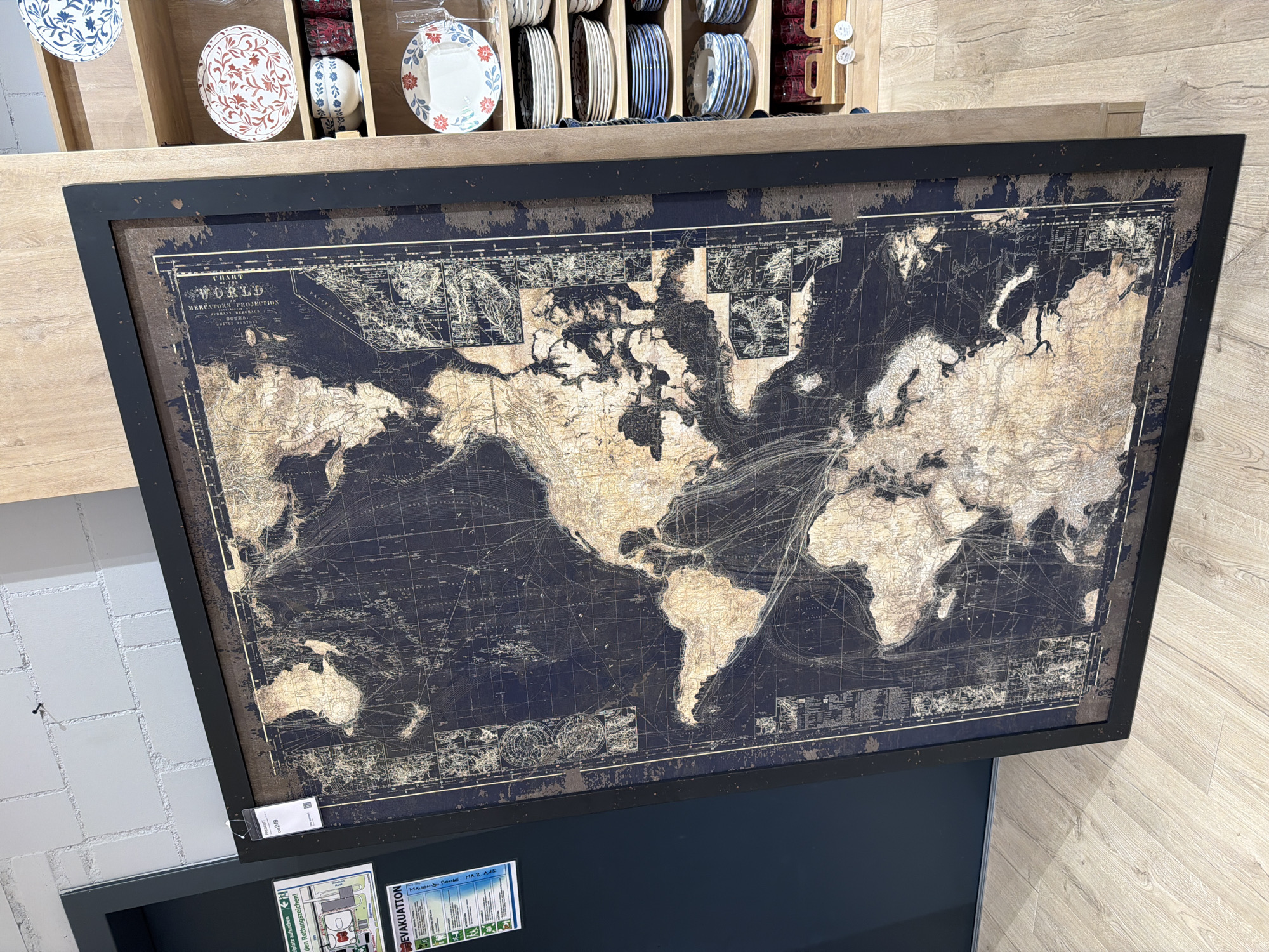 A large, framed world map is displayed prominently in what appears to be a retail setting or storage area. The map features a dark blue background with a faded, sepia-toned depiction of continents and geographical features. Overlaying the continents are intricate lines, possibly representing shipping routes or historical trade paths. The frame is black and substantial.
To the left of the map, a dark blue wall is visible, partially obscured by the map. Attached to this wall are two rectangular signs. The upper sign is white with green text and graphics, appearing to be a safety or evacuation notice, with symbols of people and a building. Below it, another sign, predominantly white with blue and red text, also seems to be related to safety or emergency procedures, with the word "EVAKUATION" prominently displayed. A price tag with "PRESCOTT" and a price of "249" is attached to the top left corner of the map's frame.
To the right of the map, a wooden shelving unit is visible. This unit is stocked with various plates and glasses. The plates are stacked in several rows, with some featuring decorative patterns in blue and red. There are also glass tumblers, some appearing to be red, stored in wooden crates on a lower shelf. The overall environment suggests a commercial space, possibly a shop or a warehouse. The floor is made of light-colored wood planks. The lighting appears to be artificial, contributing to the indoor ambiance. No people are present in the image.
