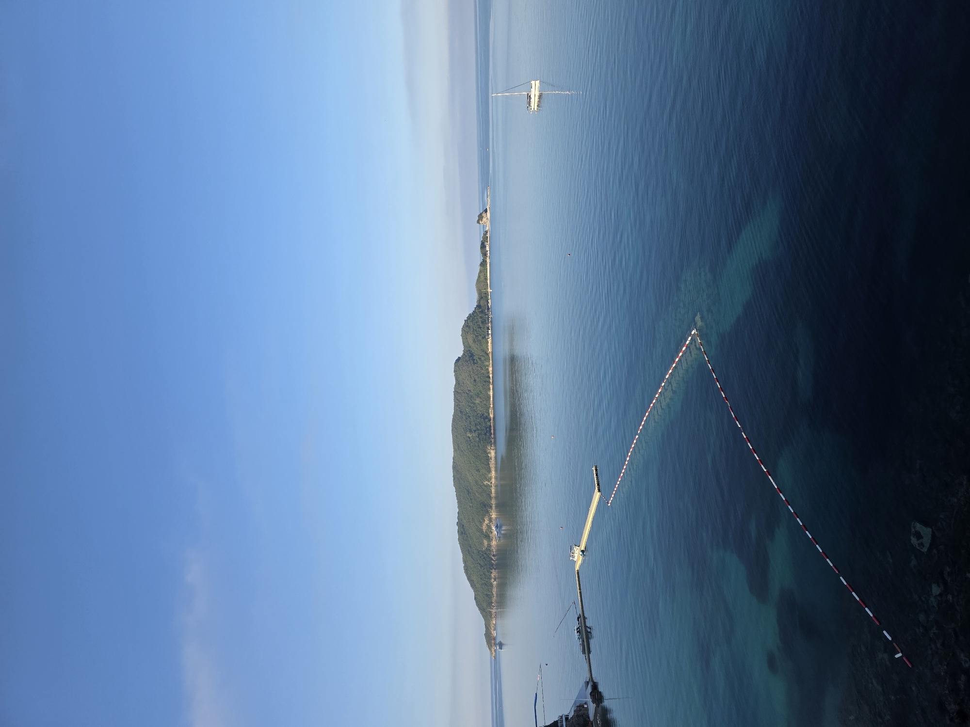 This image depicts a serene coastal scene in Budva, Montenegro, likely from an elevated viewpoint. In the foreground, the calm, clear blue water of the Adriatic Sea is visible, revealing the seabed in shallower areas. A red and white buoy line extends diagonally across the water, marking a swimming area or boundary.
A yellow floating pontoon pier extends into the sea, with a small white boat moored to it. To the left, part of a building complex or resort structure is visible, with some infrastructure such as railings and perhaps a walkway.
In the mid-ground, the sea is a deeper blue. A single white sailboat with its sail lowered is anchored in the water. Further in the distance, a large, verdant island dominates the horizon. The island is covered in dense green foliage and appears to be undeveloped. Other smaller islands or rocky outcrops are visible to the right of the main island. Several larger vessels, possibly yachts or ferries, are seen near the shore of the main island. The sky above is a clear, bright blue with a few wispy white clouds. The overall impression is one of tranquility and natural beauty, characteristic of the Montenegrin coast.