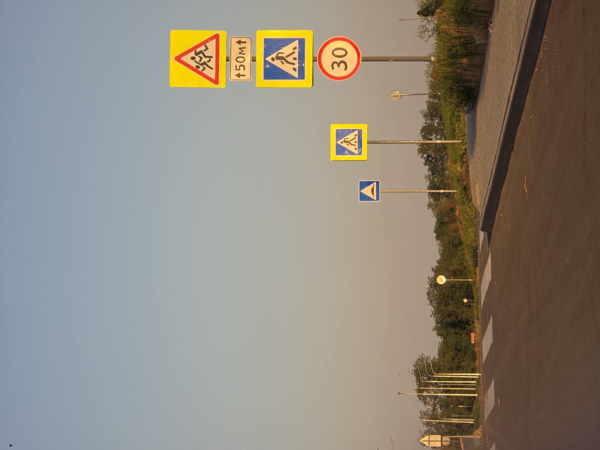 The image depicts a pedestrian crossing with multiple road signs in the city of Rostov-on-Don, Russia. The scene is characterized by a clear sky with a gradient of light blue to pale orange, indicating either early morning or late afternoon. The foreground shows a freshly paved asphalt road with a white striped pedestrian crossing. To the right of the crossing, a concrete sidewalk is visible, bordered by overgrown grass and bushes. A series of road signs are prominently displayed on poles. These include a triangular sign with a silhouette of running children, a rectangular sign indicating "150m" with an upward arrow, a square blue sign with a white pedestrian silhouette, and a circular red sign with the number "30", indicating a speed limit. Further in the background, several streetlights and trees line the road. The overall impression is one of a newly developed or maintained area, likely designed to improve pedestrian safety and traffic management.